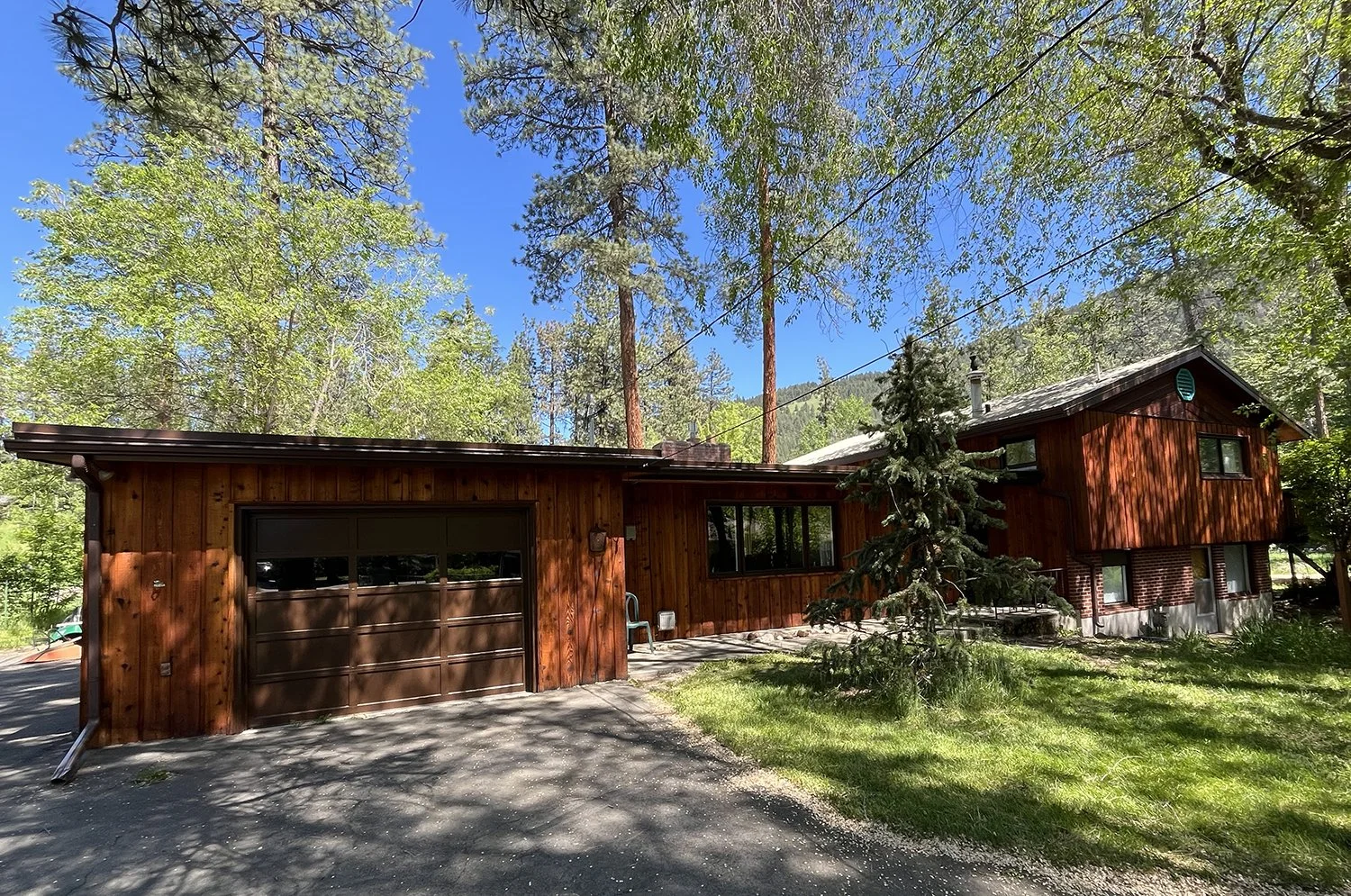 A wooden house surrounded by tall trees, with a garage door in the front and windows reflecting the sky and trees, in a lush green setting with a clear blue sky.