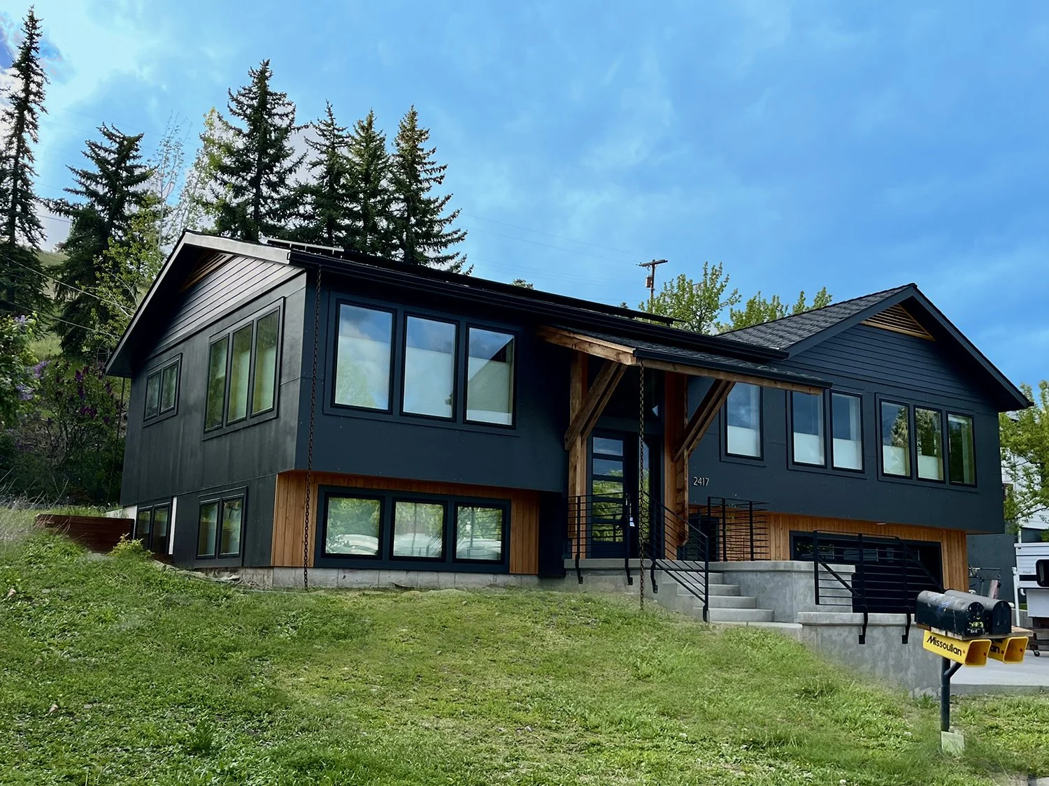 Modern two-story house with black siding and large windows, set on a grassy lot with tall trees in the background, featuring a concrete staircase and a black railing leading to the front entrance.