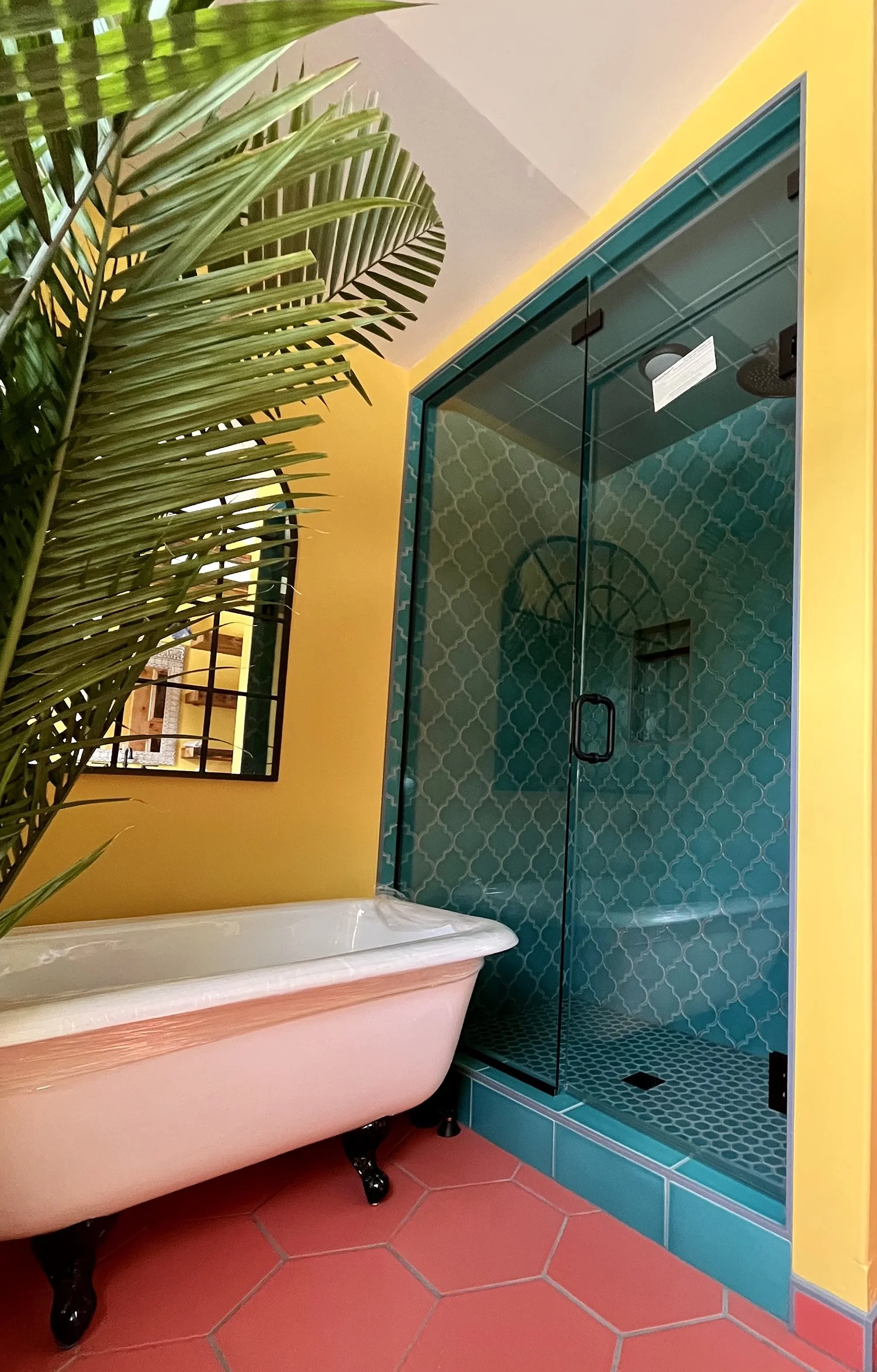 A colorful bathroom featuring a clawfoot bathtub, a glass shower enclosure with blue patterned tiles, yellow walls, a hexagonal terracotta tile floor, a mirror, and a large green plant.