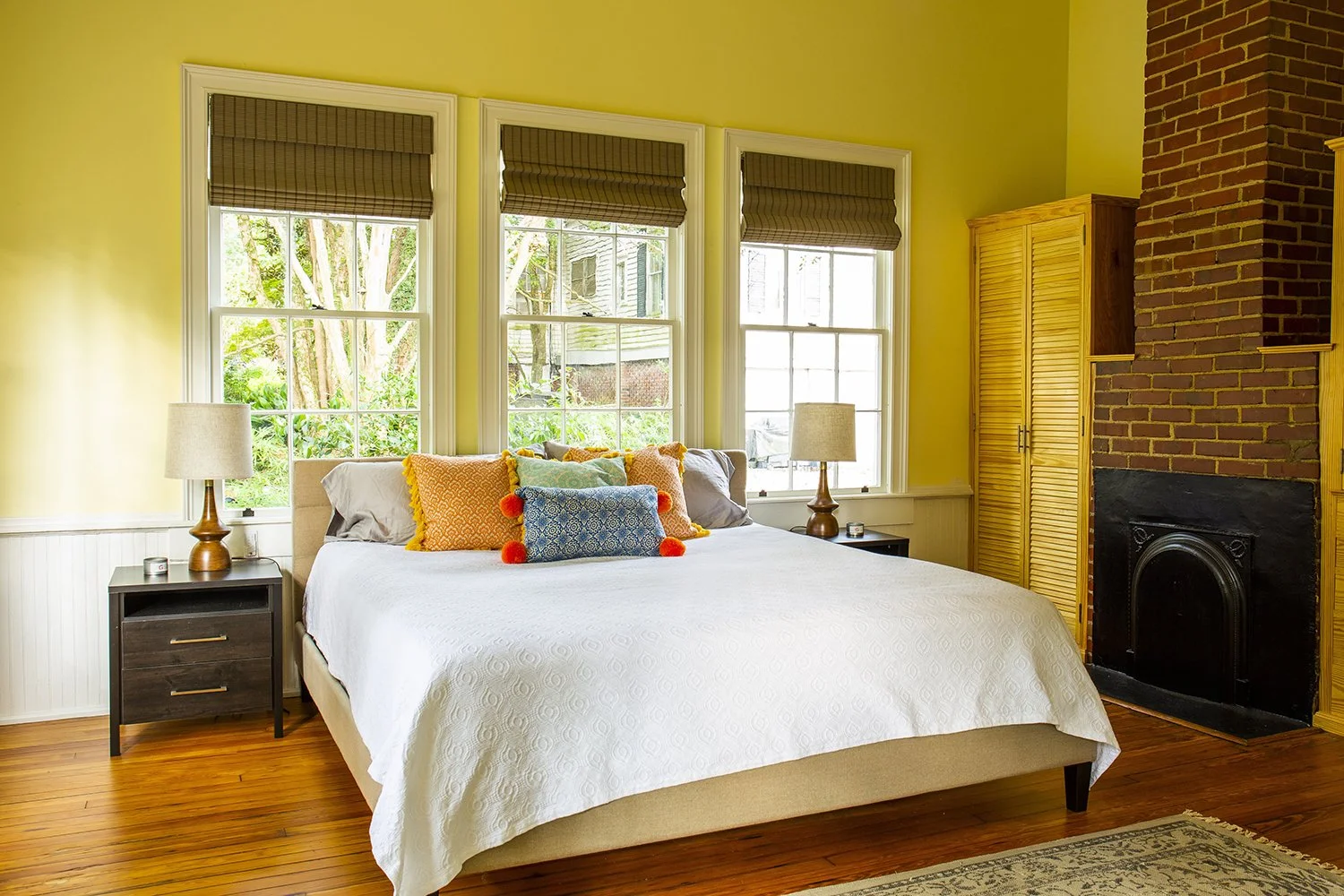 Bright bedroom with a large bed, orange, blue, and gray pillows, two black nightstands with white lamps, three windows with brown blinds, yellow walls, wooden floors, and a brick fireplace with wooden cabinets.