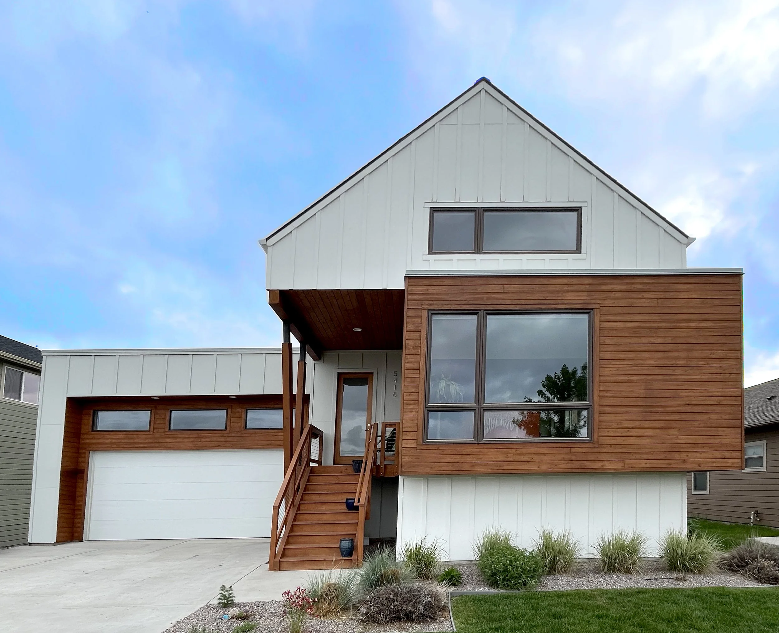 Modern two-story house with a white and wood exterior, a garage, and a front porch with stairs. Landscaping includes small shrubs and grass.