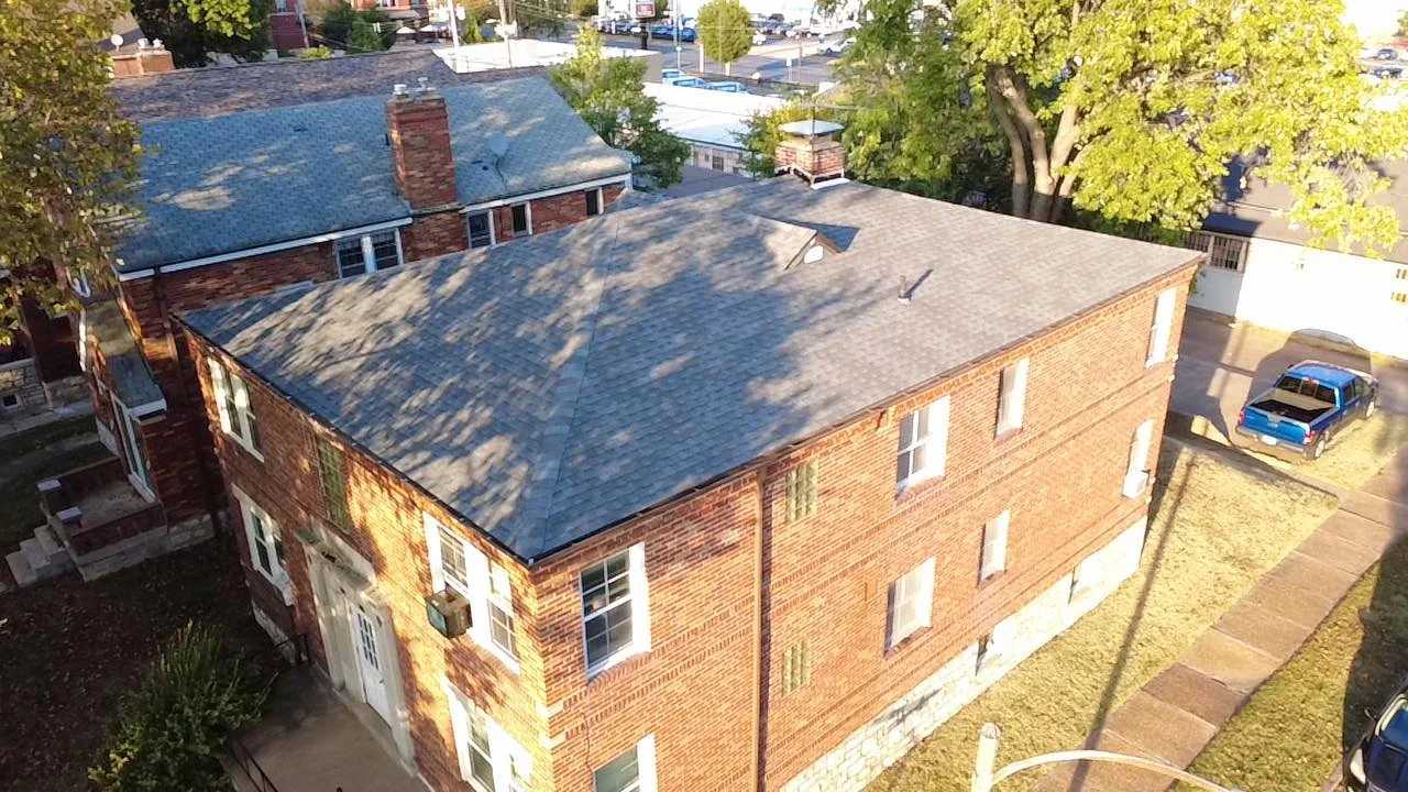 An aerial view of a brick two-story commercial apartment building with a gray shingle roof by National Roofing St. Louis, surrounded by trees and a driveway with parked cars.