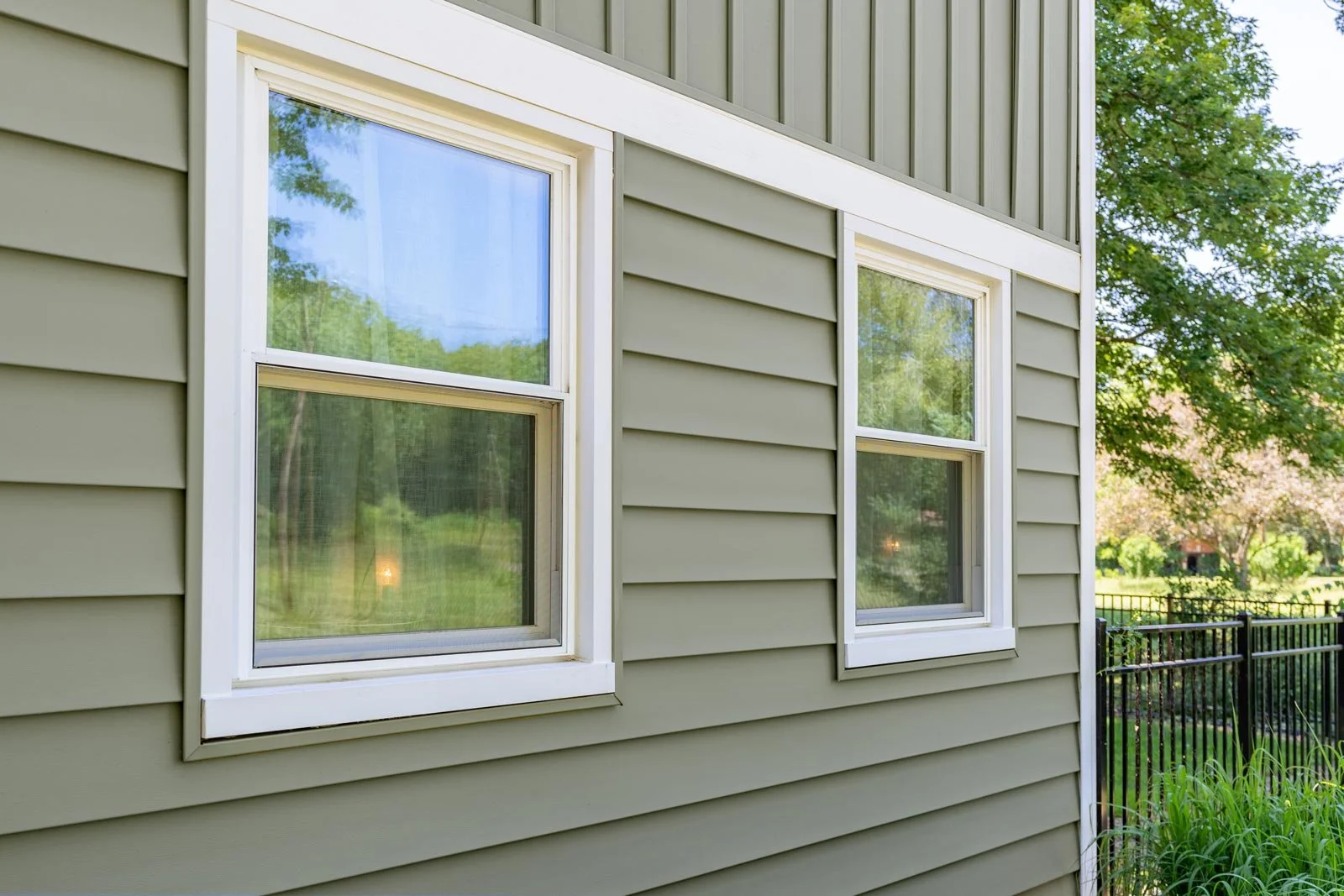 Exterior of a house with olive green vinyl siding by National Roofing St. Louis and three double-hung windows with white frames, showing a lush green yard and trees outside.