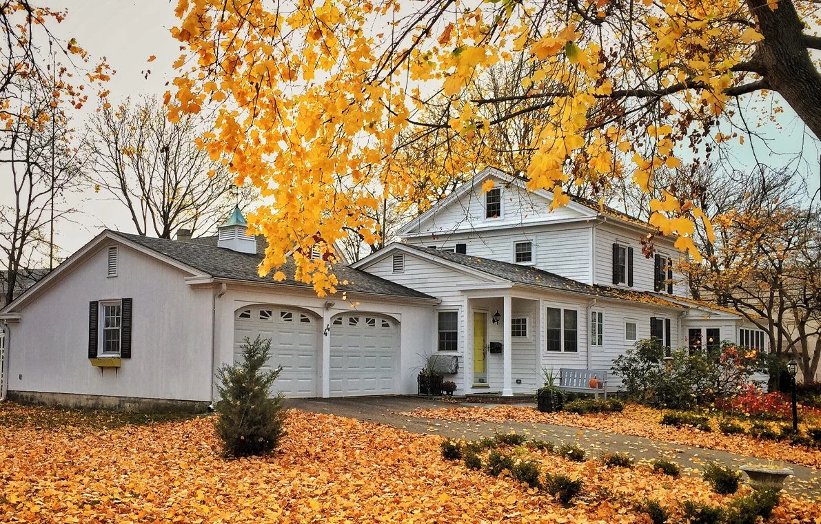 A white house with black shutters and a yellow front door, surrounded by trees with yellow leaves in autumn.