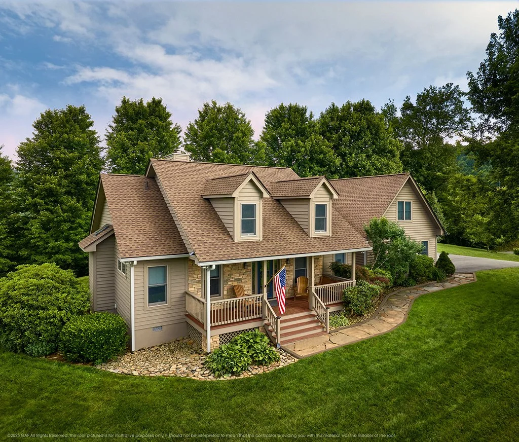A house with a brown shingled roof by National Roofing St. Louis and beige siding, a front porch with Adirondack chairs, a flag, and surrounded by green trees and grass.