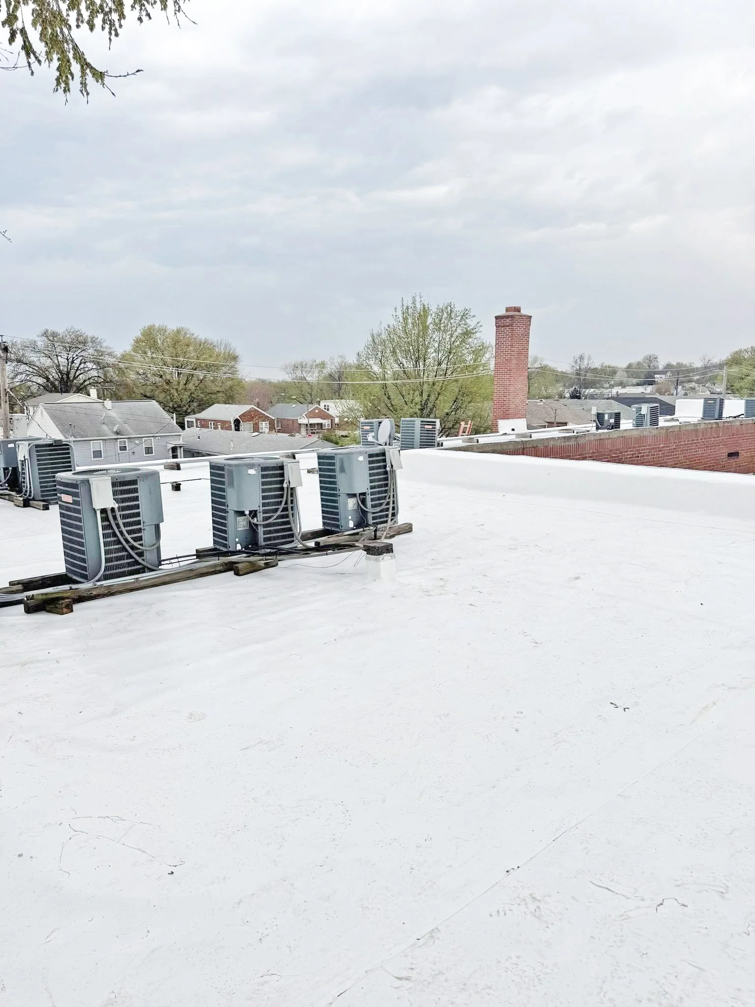 View of a flat white commercial rooftop by National Roofing St. Louis with several air conditioning units installed on metal brackets. In the background, there are neighborhood houses with trees and a red brick chimney visible under a cloudy sky.