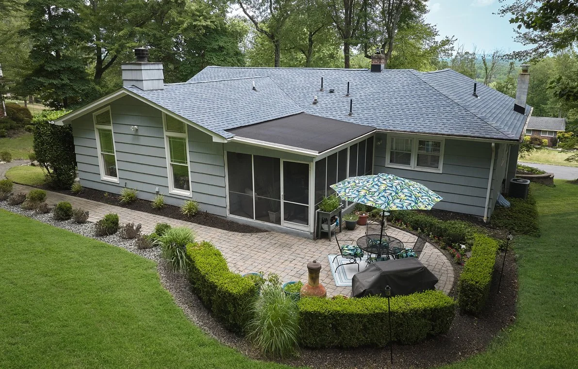 Backyard patio with table, chairs, umbrella, and grill, enclosed by a screened porch and manicured lawn with landscaped bushes and trees surrounding a light blue house with a vinyl siding exterior.