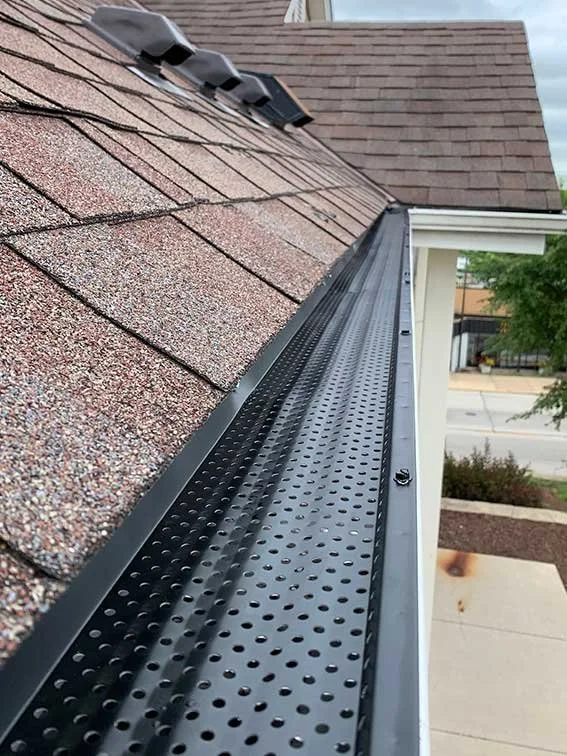 Close-up of a new black perforated metal gutter attached along the edge of a shingled roof by National Roofing St. Louis to protect home roof from leaks and storm damage.