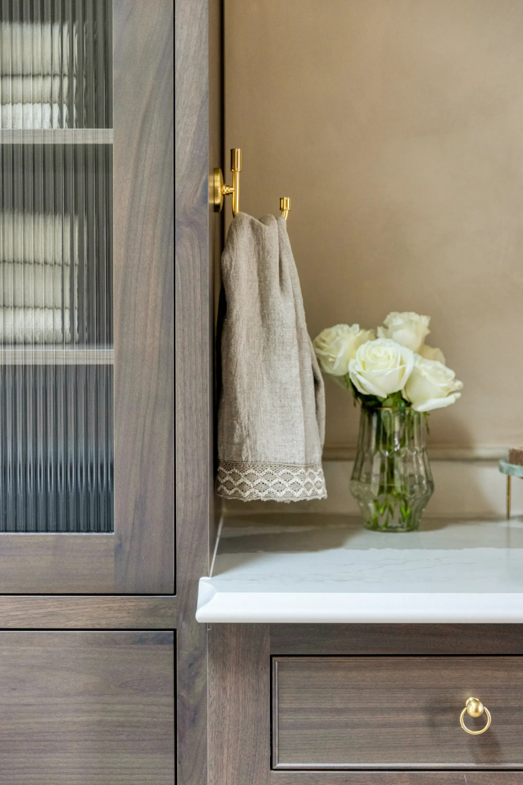 S. Flynn Design + Build Interior Design showcasing wooded cabinets, gold hardware, beige linen towels and a green vase with white flowers on a marble sink.