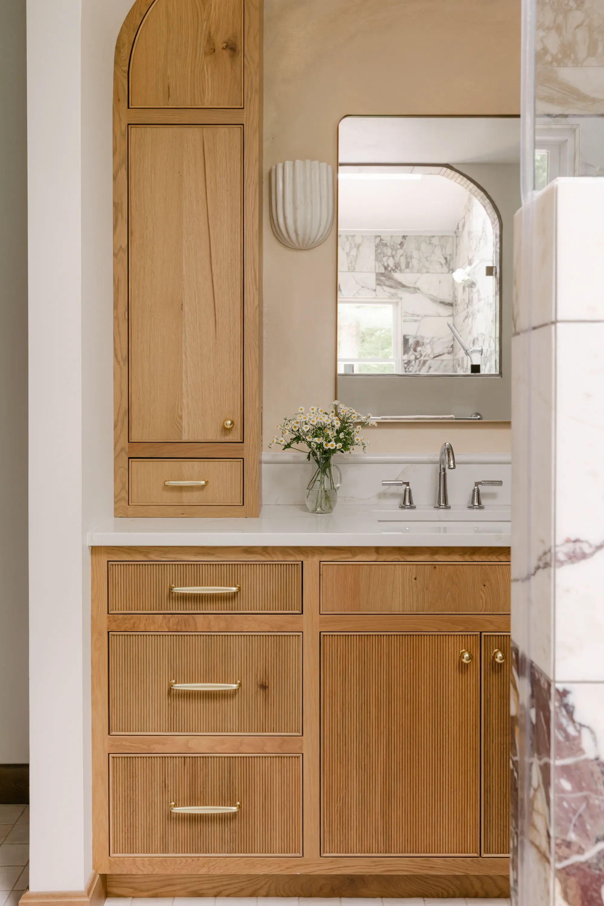 S. Flynn Design + Build interior design for bathroom remodel showcasing warm brown wood cabinets, a marble counter top, gold hardware, and modern curved mirror.