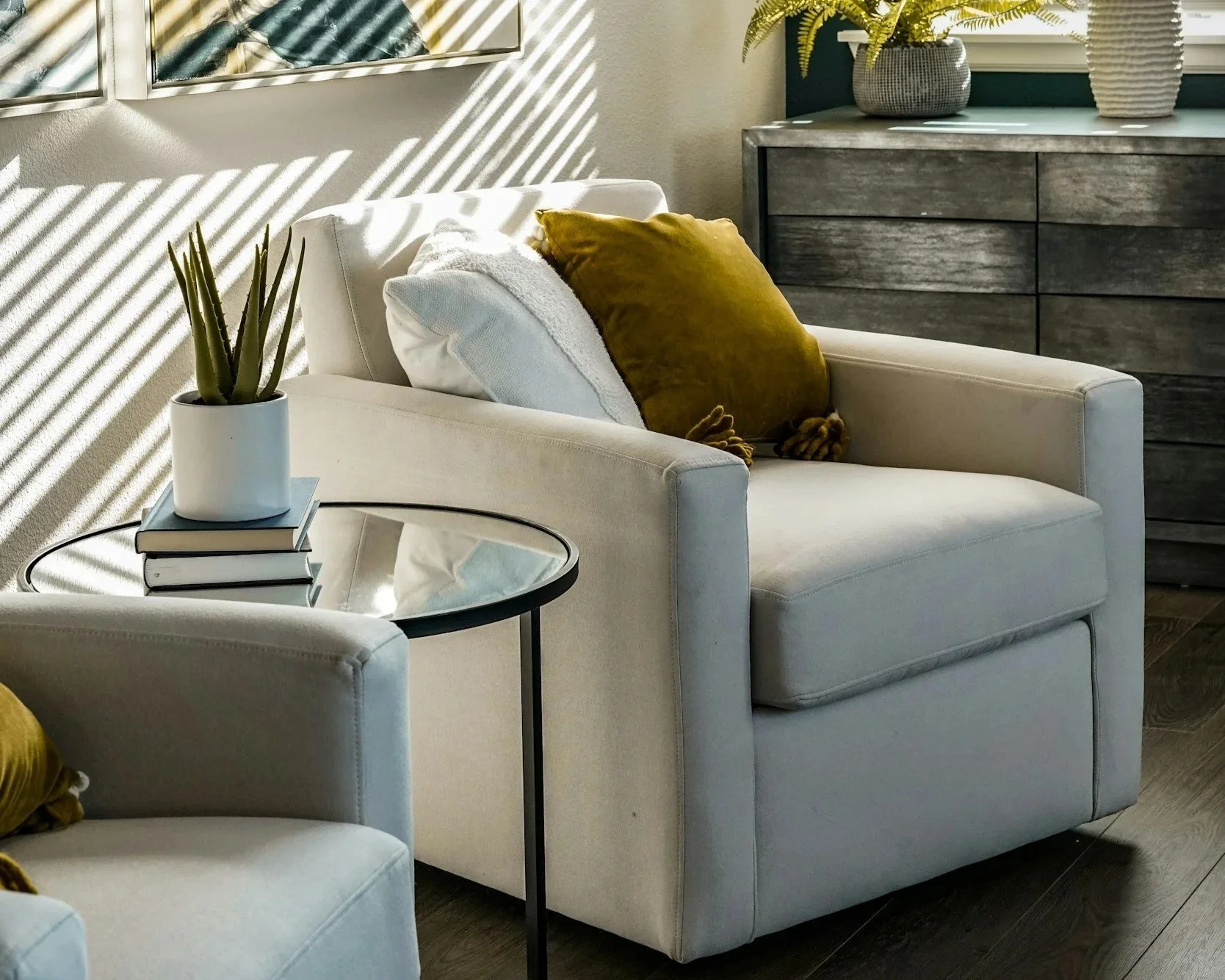 A cozy living room corner with a beige chair, a glass-topped side table with a potted succulent and stacked books, and a dark wooden dresser topped with potted plants. Sunlight filters through window blinds, creating striped shadows on the wall.