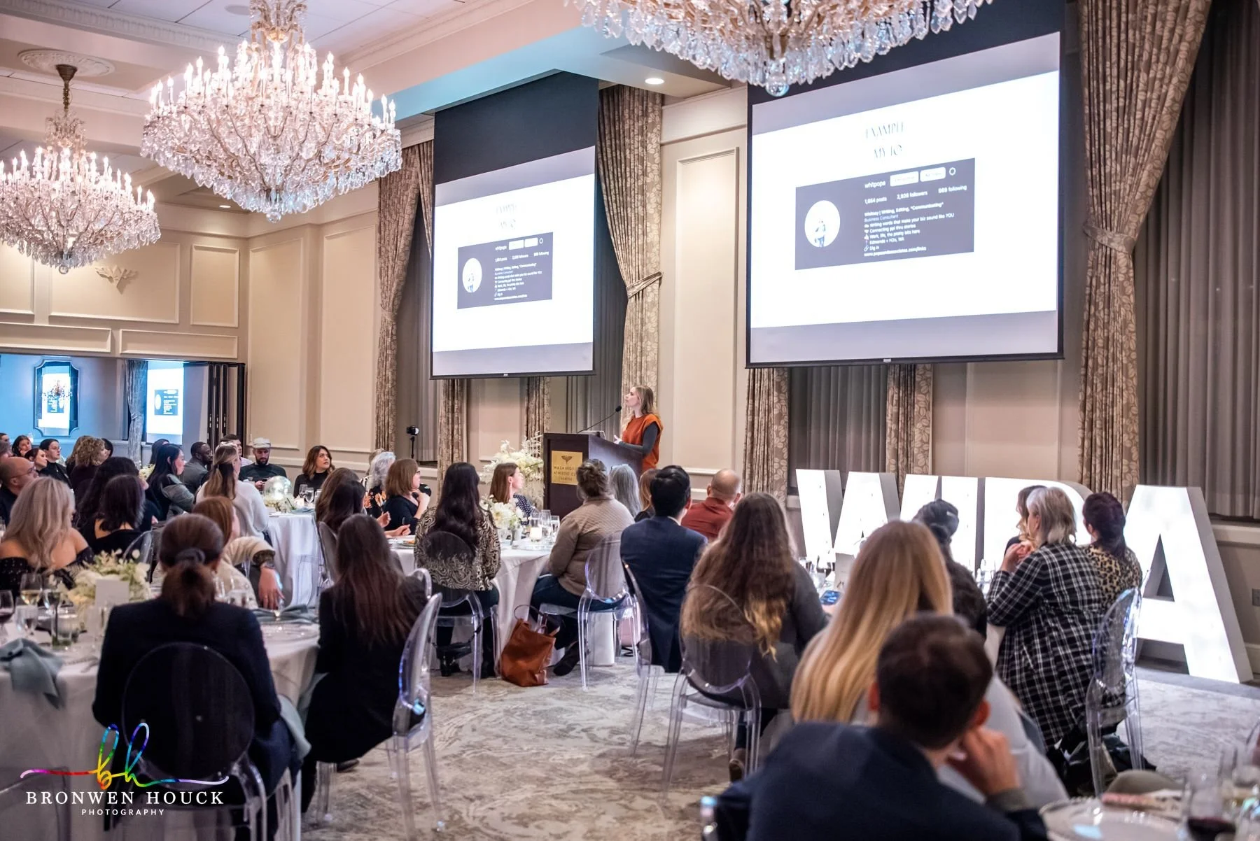 A large formal event or conference with many people seated at round tables, listening to a woman speaker at a podium in a grand, elegantly decorated ballroom with chandeliers and large screens displaying presentation slides.