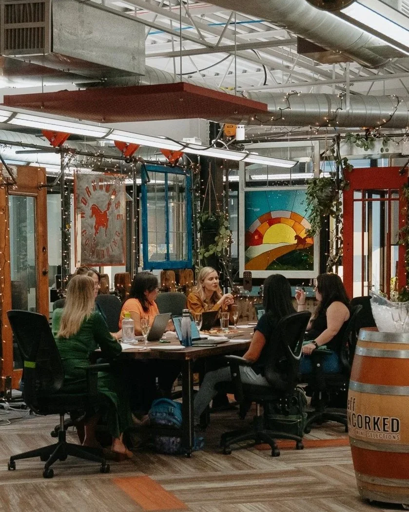 Six women sitting around a rectangular table in an industrial-style office or co-working space, engaged in conversation. The space features exposed ducts and pipes, festive string lights, colorful abstract artwork, and some decorative plants.