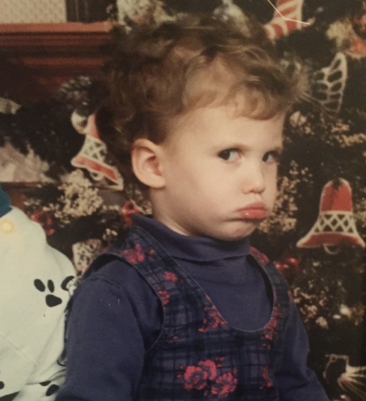 A young child with curly hair and a serious expression standing in front of a decorated Christmas tree, wearing a blue shirt and plaid vest with floral embroidery.