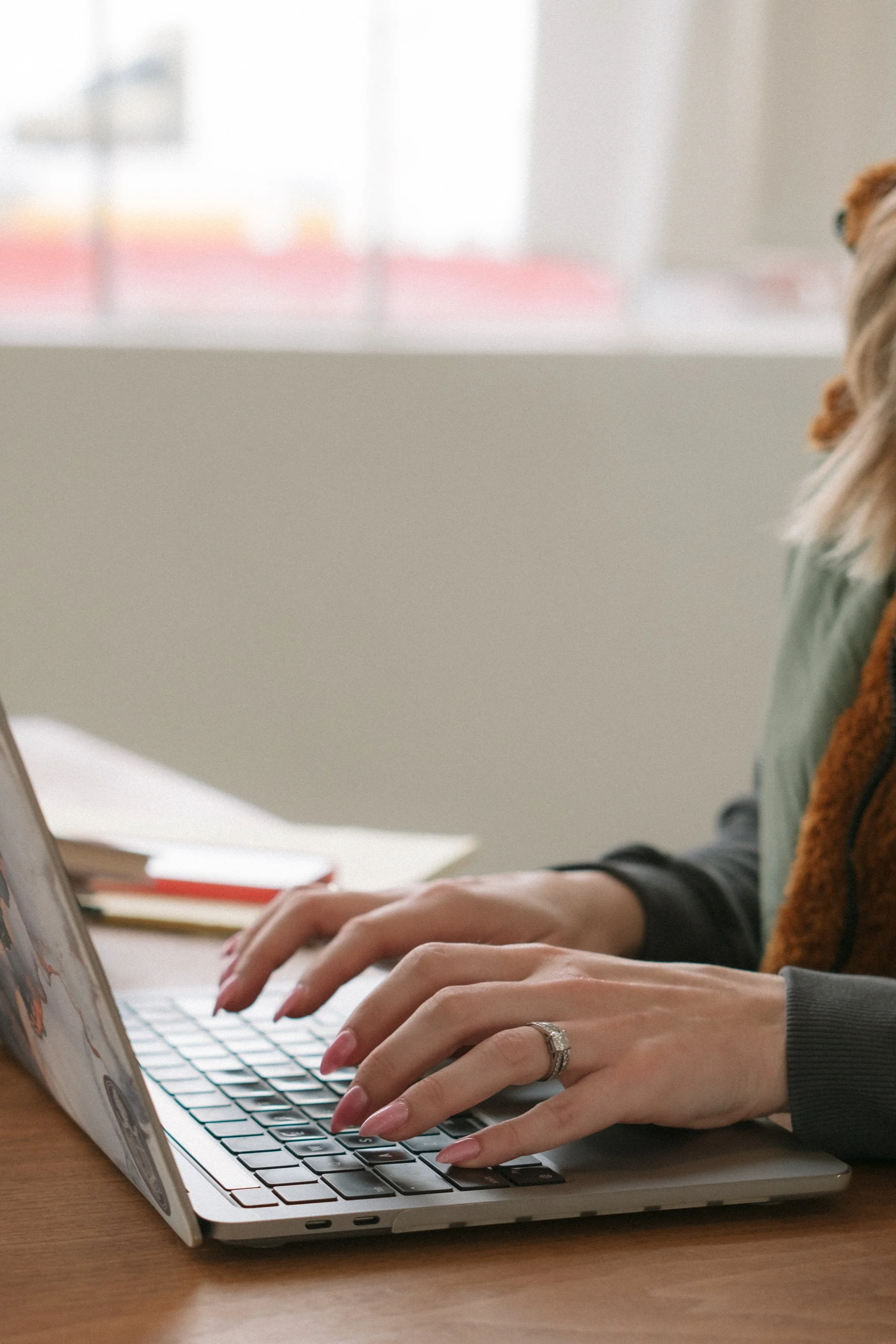 Close-up of a person typing on a laptop keyboard at a wooden desk, wearing a ring on their left hand, with a blurred background of a window and some books.