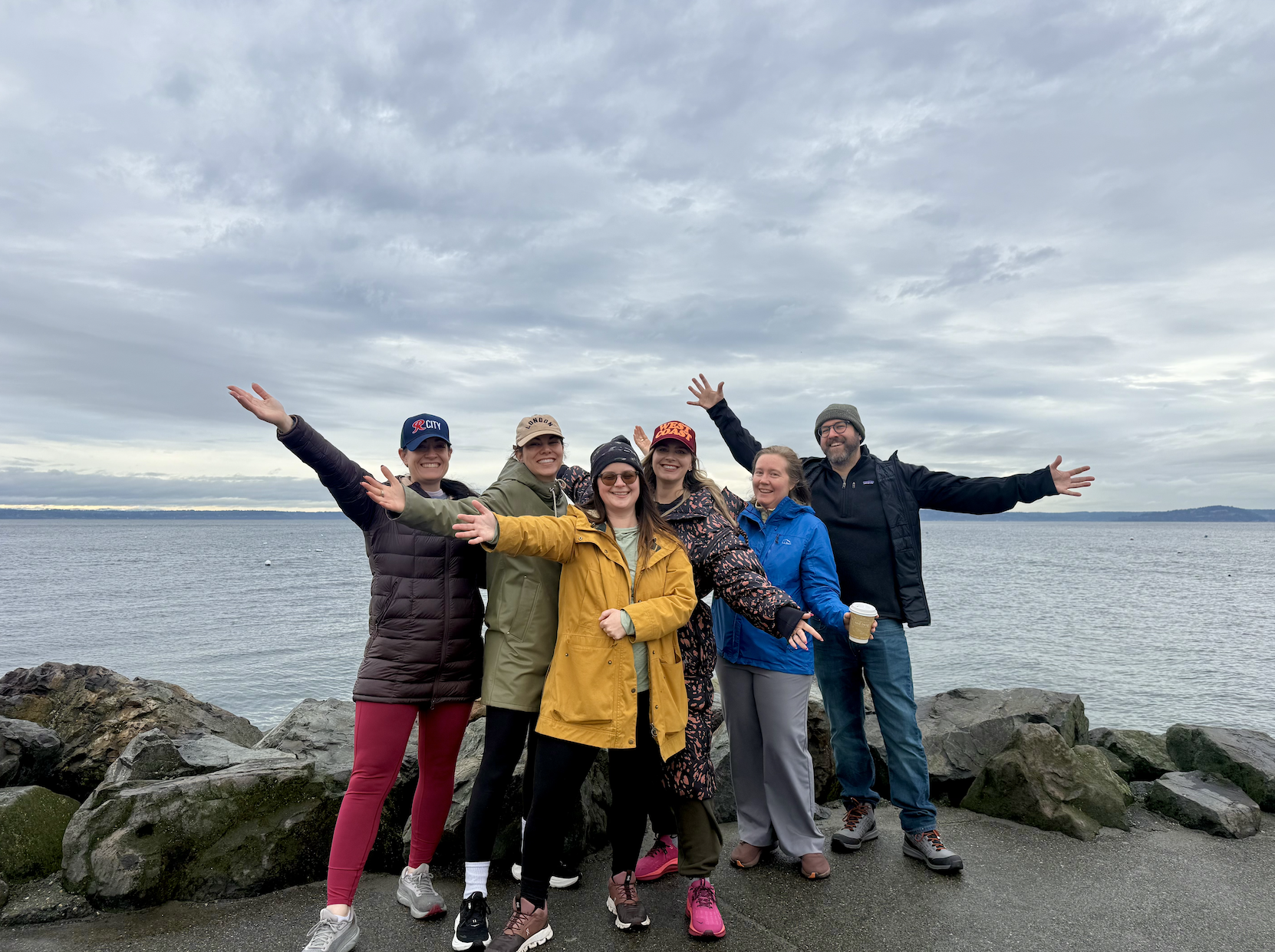 Group of six people standing on rocks by a body of water with a cloudy sky, smiling and raising their arms in celebration.