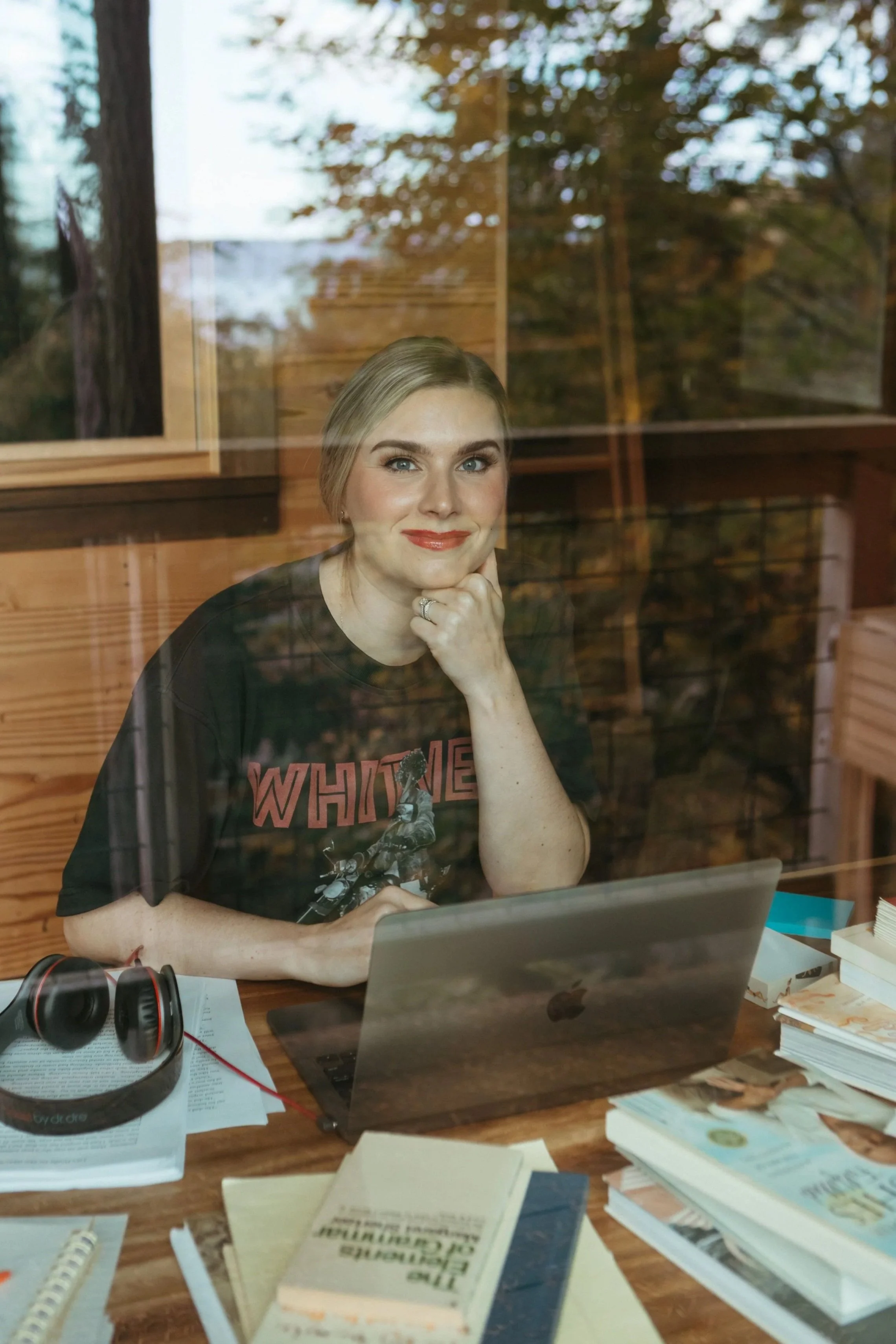 A woman with blonde hair and blue eyes sits at a wooden desk, smiling and resting her chin on her hand, in front of a laptop with a glass window behind her showing trees outside. The desk has notebooks, books, papers, a pair of headphones, and a pen.