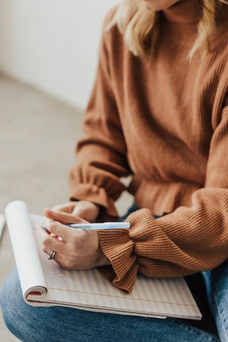 A person wearing a brown long sleeve shirt and blue jeans, sitting and writing in a notebook with a pen, with a rolled-up piece of paper nearby.