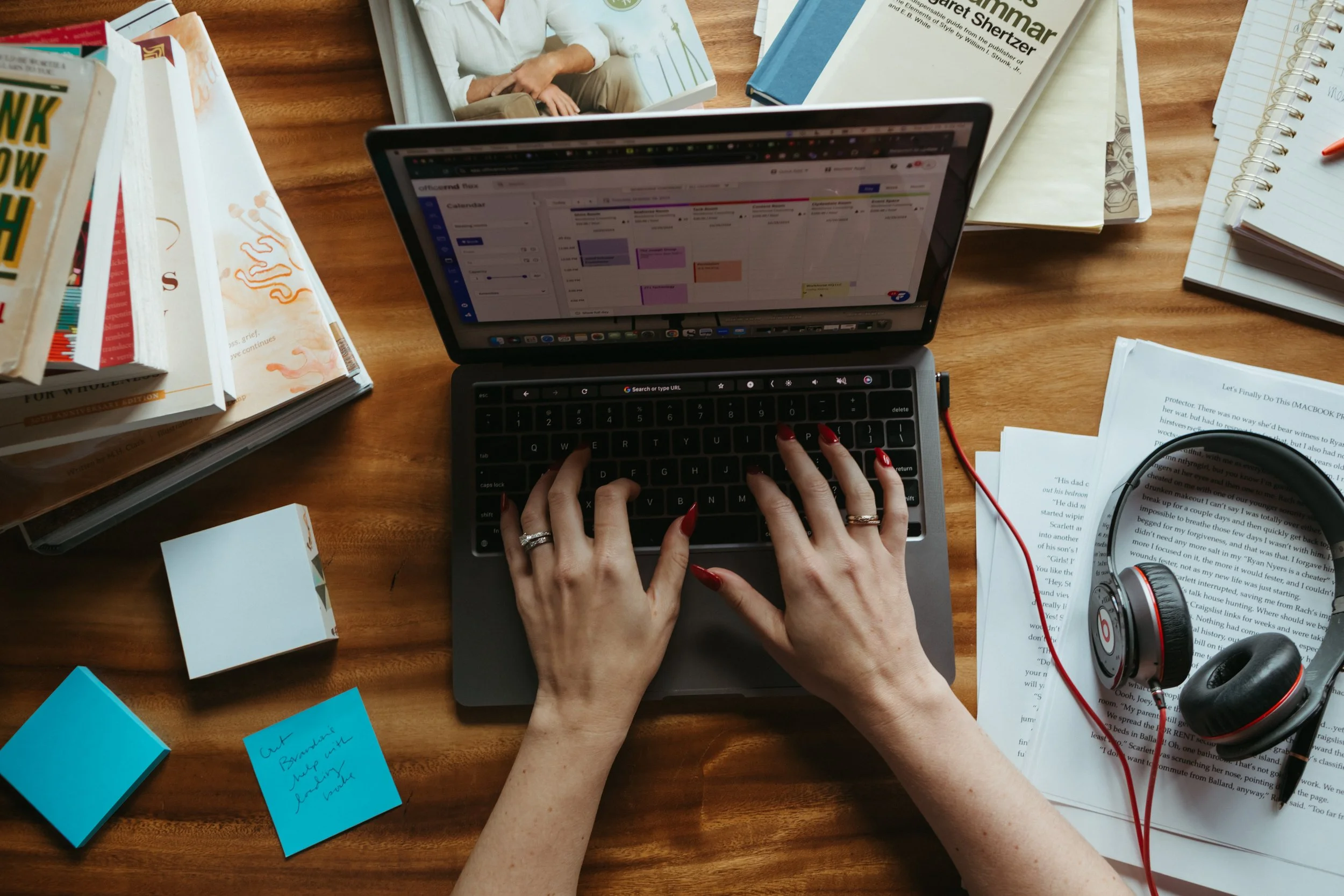 A person's hands with red nails typing on a small silver laptop on a wooden desk surrounded by books, notebooks, sticky notes, printed documents, and a pair of headphones.