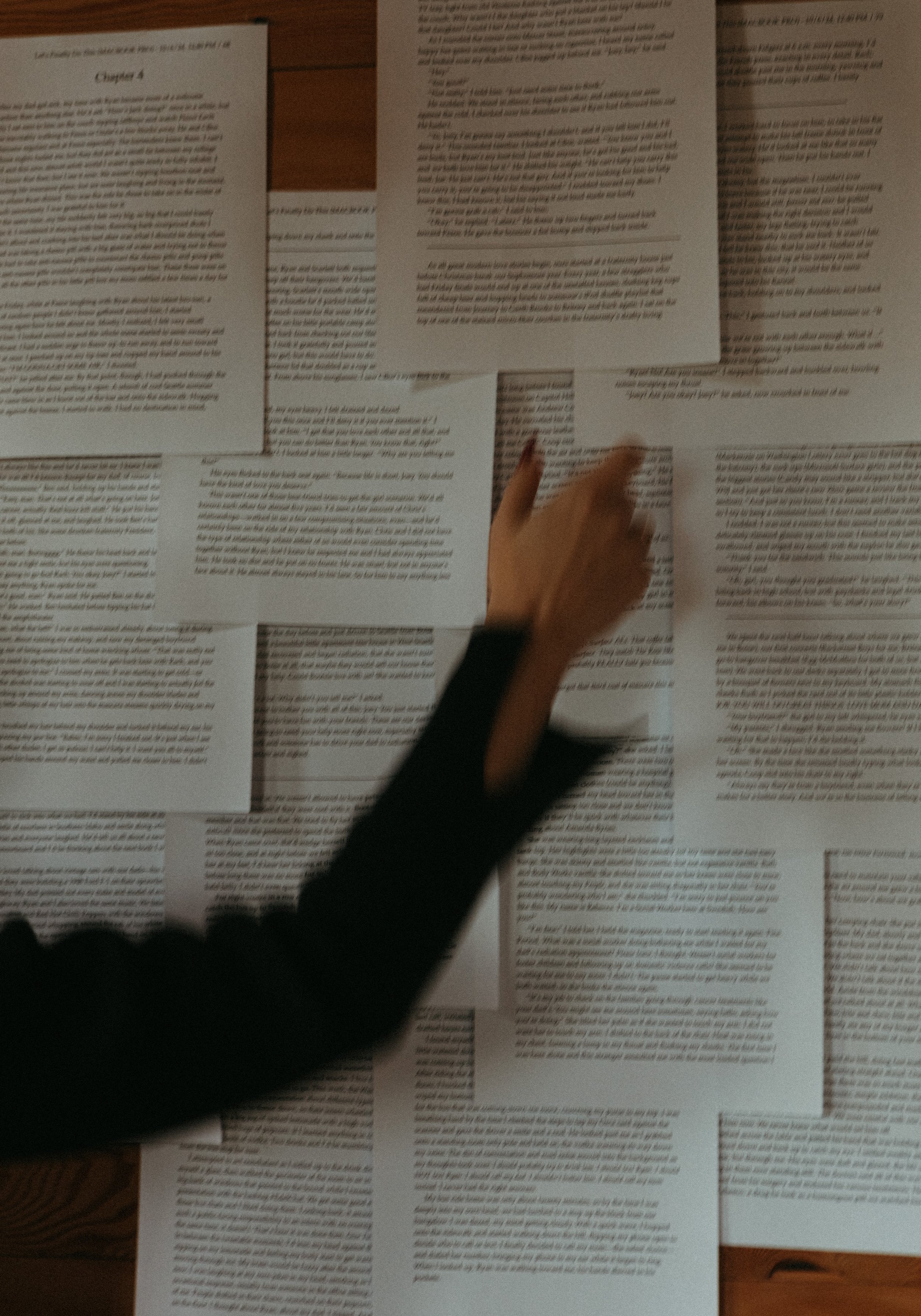 Several sheets of paper with printed text spread out on a wooden surface, with a hand reaching towards one of the sheets.