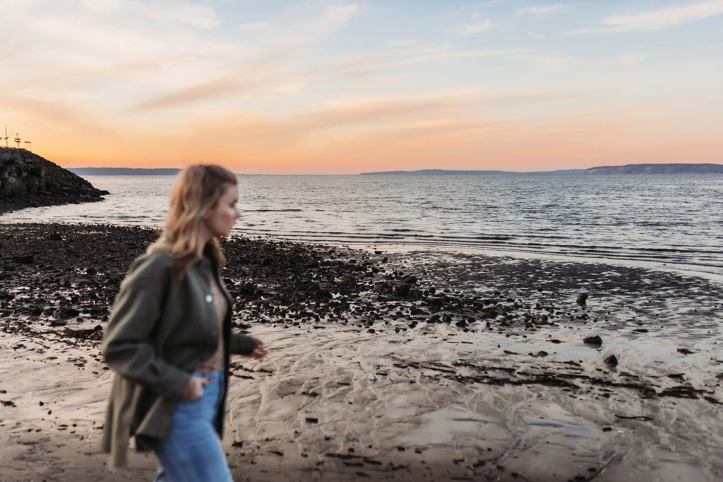 A woman with wavy hair wearing a jacket walking along a rocky beach at sunset with calm water and distant land in the background.