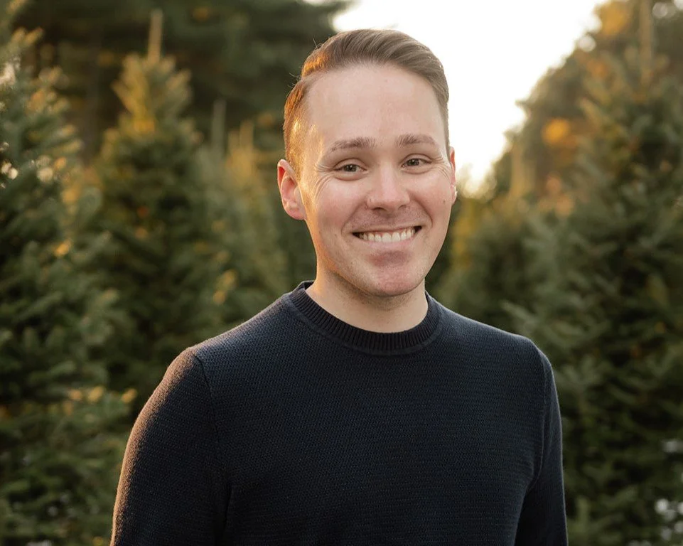 A young man with short brown hair and a wide smile, wearing a black sweater, standing outdoors among green pine trees during golden hour.