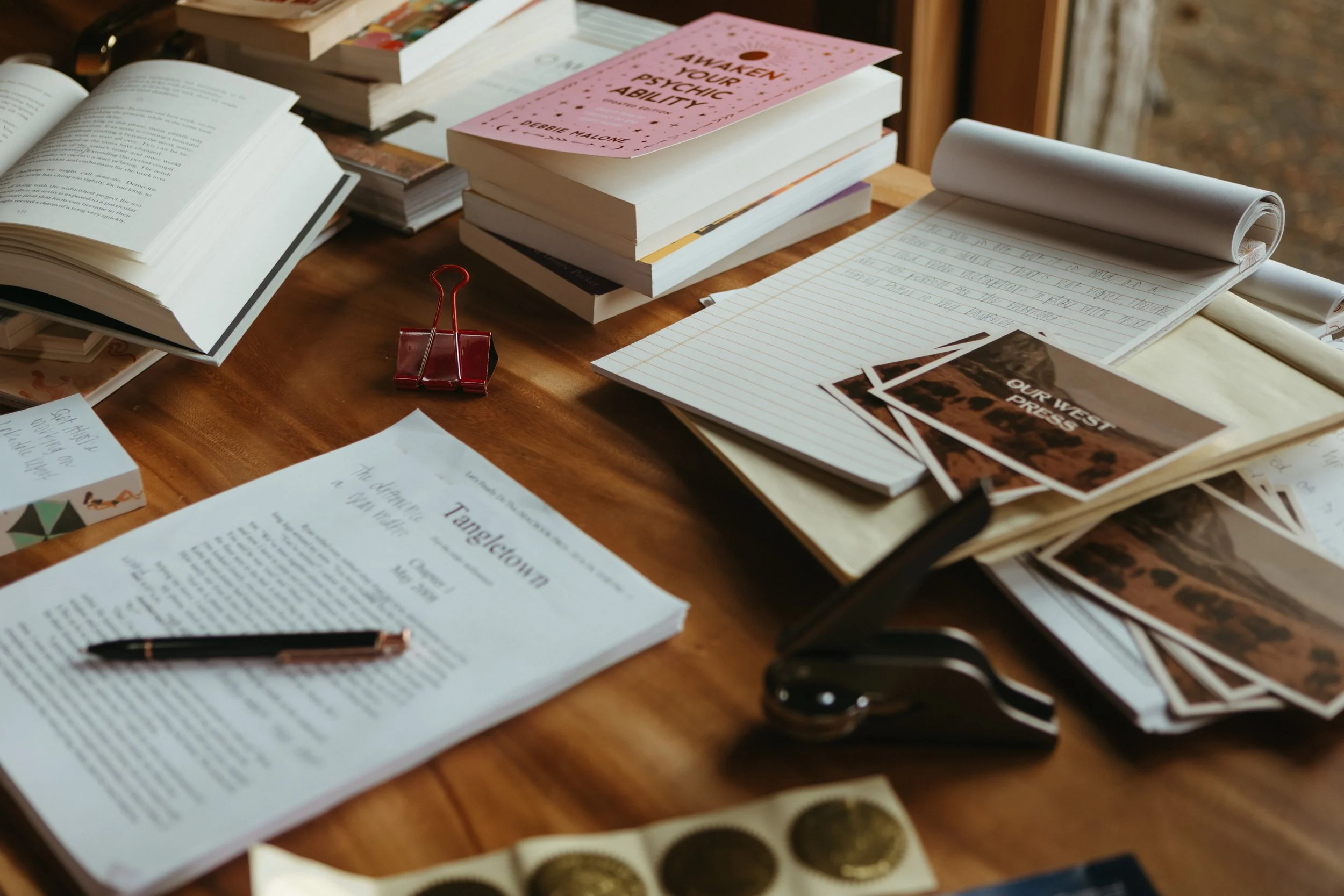 A cluttered wooden table with open books, notebooks, notes, a red binder clip, and some paper. The scene appears to be in a cozy indoor space, possibly a workspace or study area.