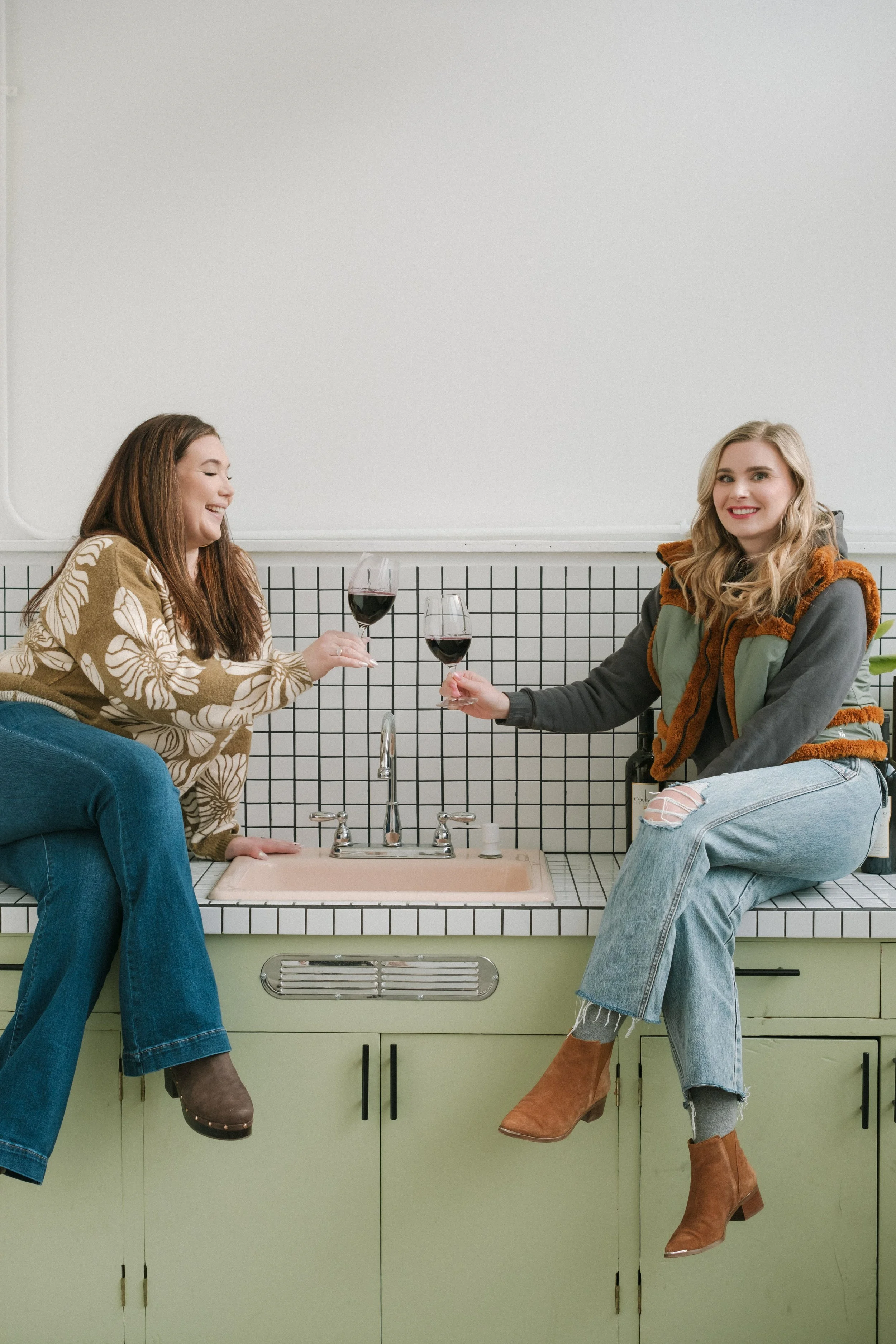 Two women sitting on a kitchen counter, holding glasses of red wine, smiling and engaging in conversation. One woman has long brown hair and is wearing a floral patterned brown sweater and blue jeans. The other woman has long blonde hair and is wearing a gray and green jacket with a brown fur collar, ripped jeans, and brown boots. The kitchen features a green cabinet, a pink sink, and a black and white tiled backsplash.