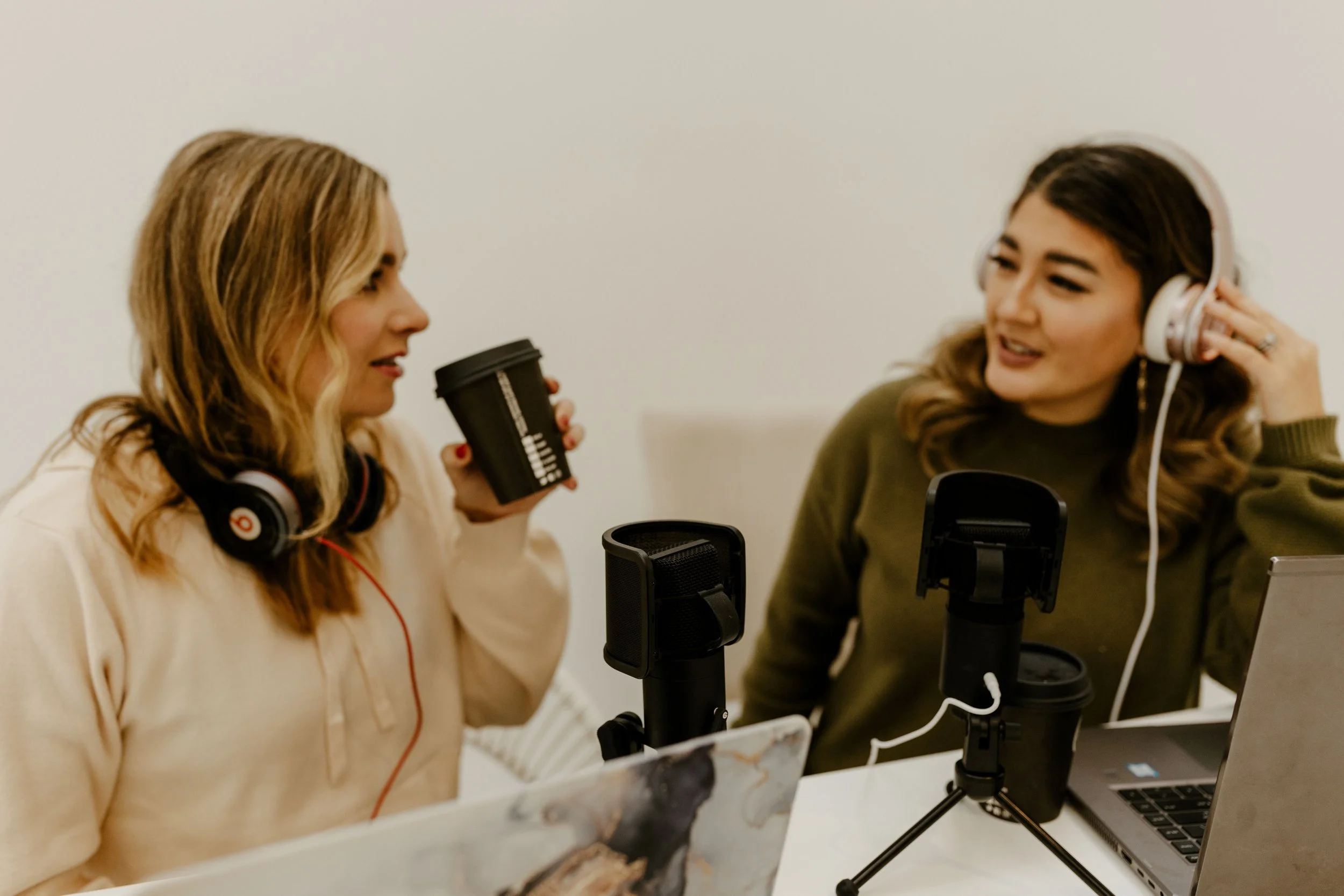 Two women with headphones engaged in a recording session, one holding a coffee cup, with a microphone and laptop on a white table.