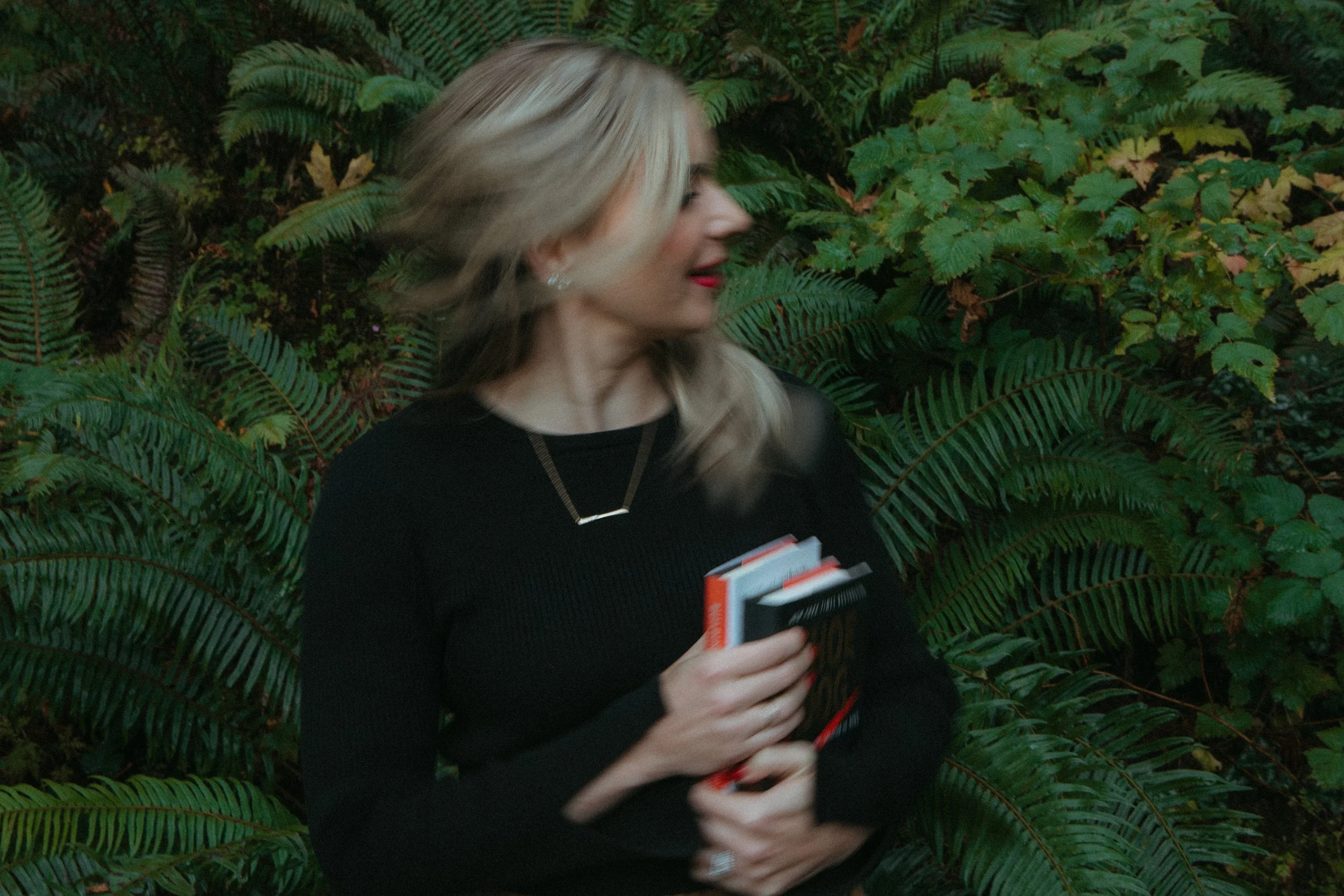 A woman with long blonde hair, wearing a black top and red lipstick, is holding books and standing among lush green fern plants.