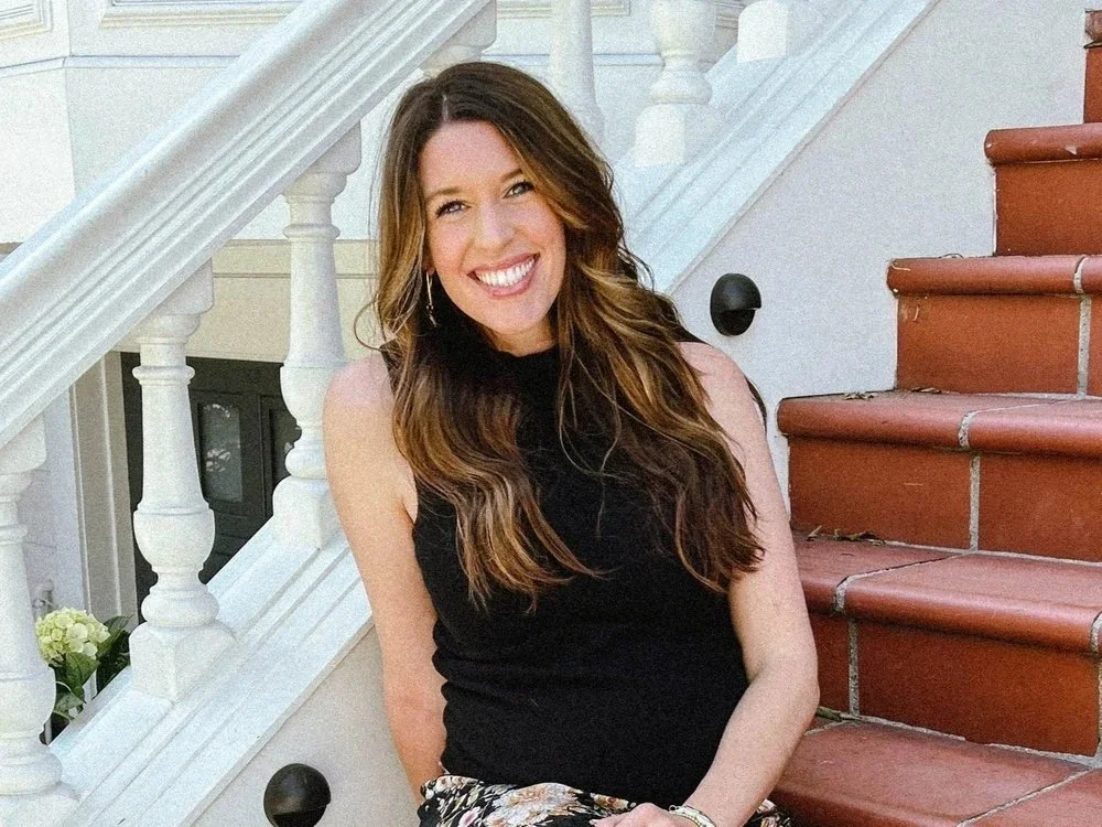 A woman with long wavy brown hair wearing a black sleeveless top, sitting on red stairs outside near a white railing, smiling at the camera.