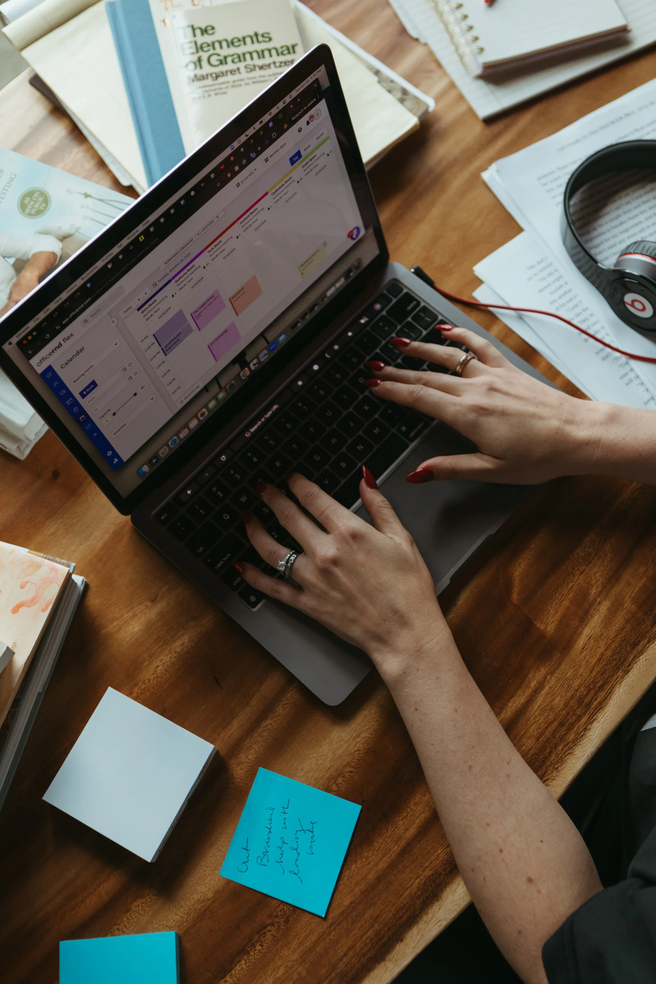 A person with painted nails using a laptop with color-coded calendar schedules on the screen, surrounded by notebooks, printed documents, colorful sticky notes, and a pair of headphones on a wooden desk.