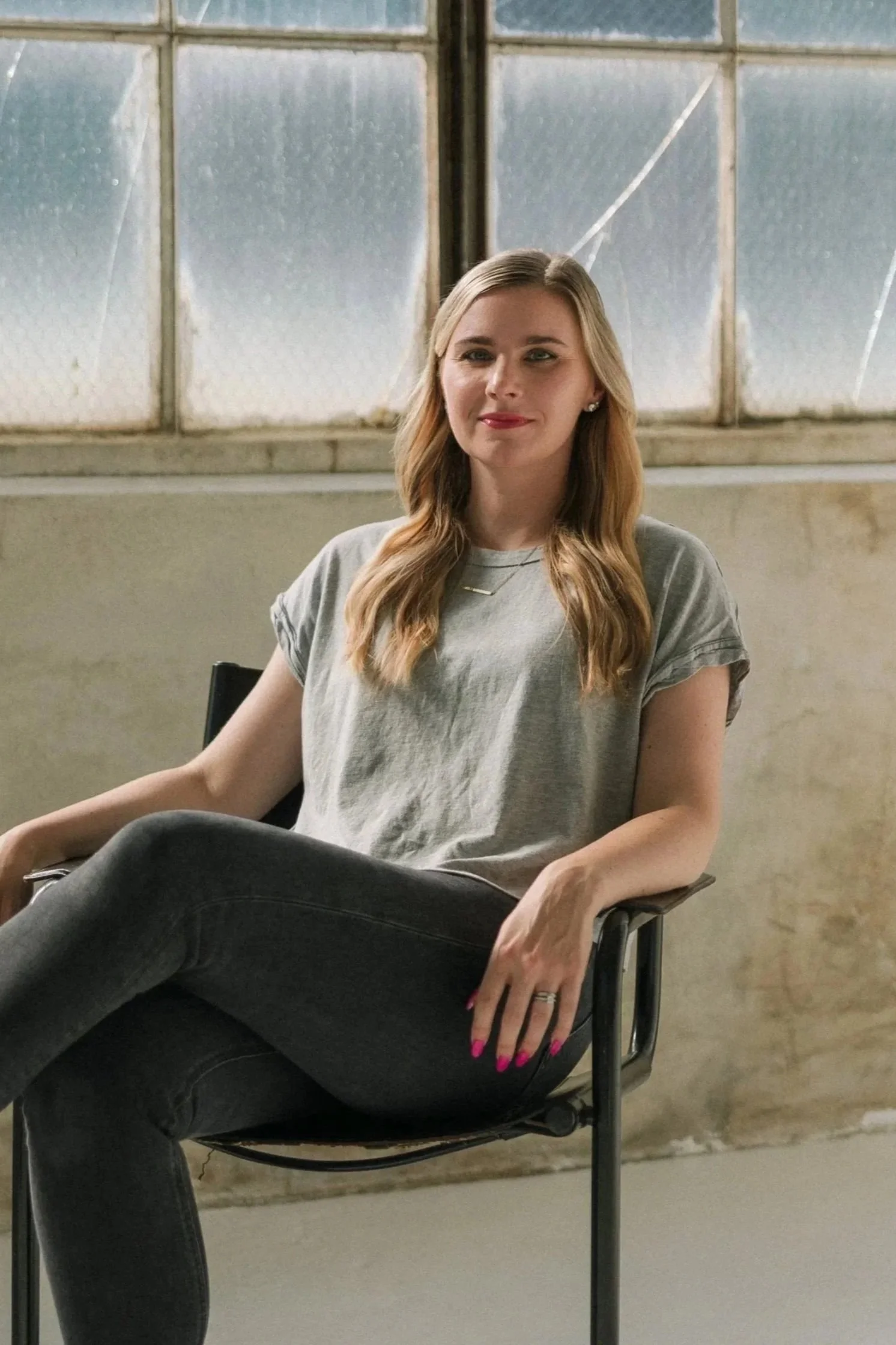 A woman with long wavy blonde hair sitting in a black chair in front of frosted window pane. She is wearing a gray T-shirt, black pants, a small necklace, pearl earrings, and has pink painted fingernails. She is smiling subtly with a relaxed posture.
