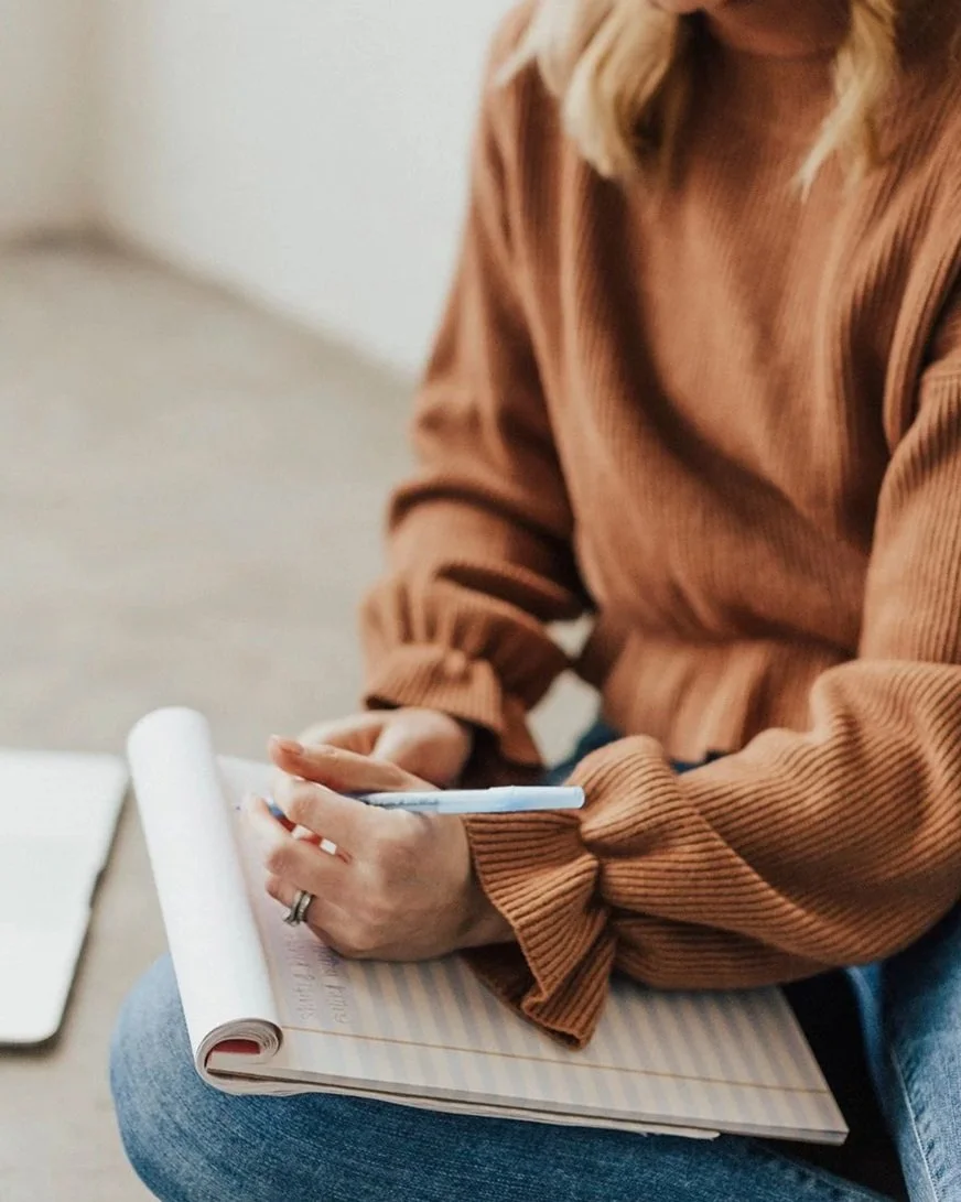 Person sitting with a notebook and pen, wearing a brown long-sleeve shirt and blue jeans.