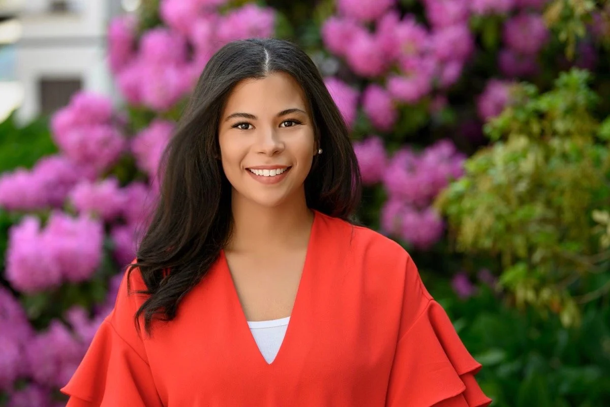 A young woman with long dark hair smiling outdoors, with pink flowering bushes in the background, wearing a red top and white undershirt.