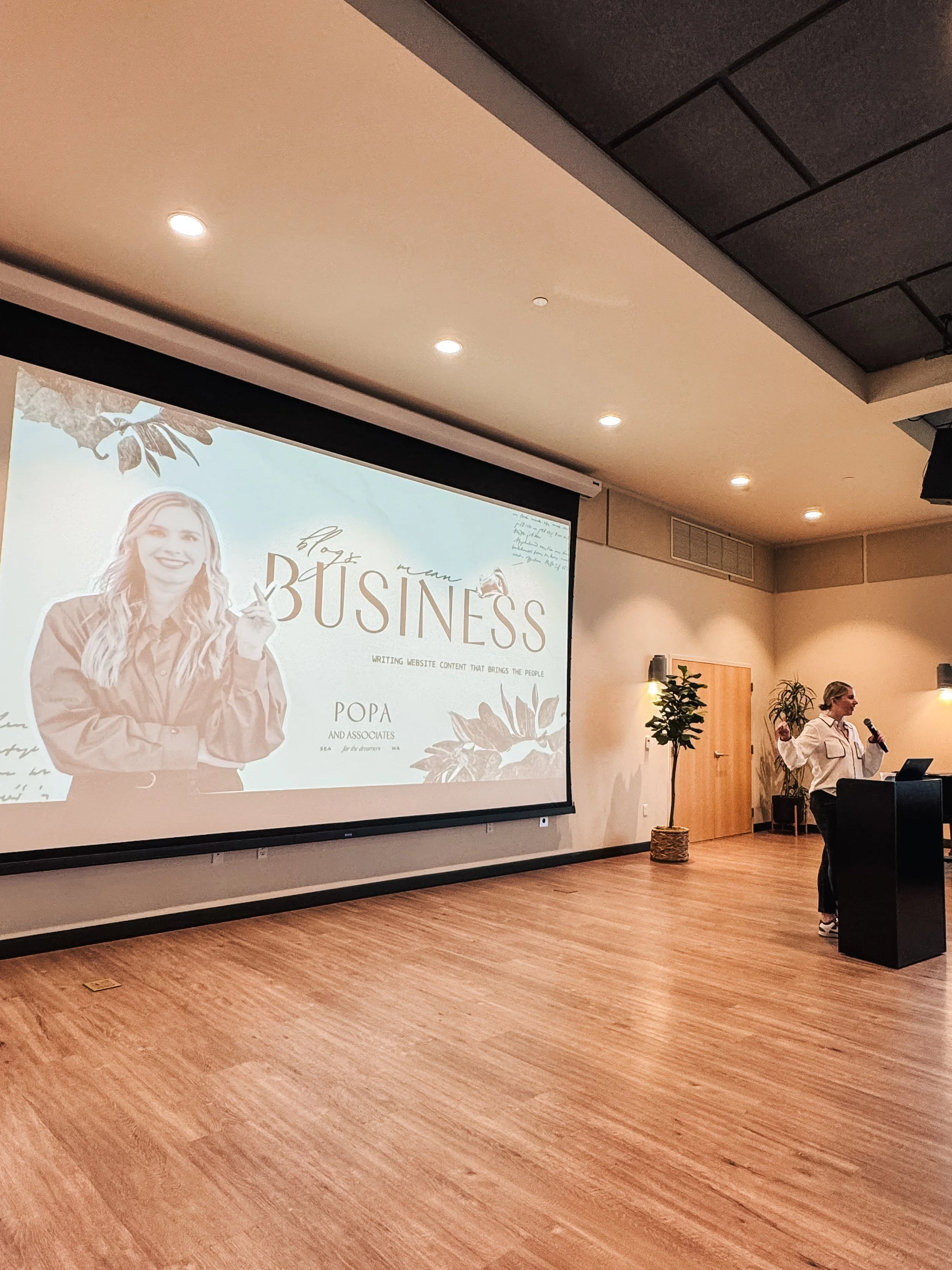 A presentation room with a projector screen displaying a slide titled 'blog mean BUSINESS' with a photo of a smiling woman, and a woman speaking at a podium with microphones, plants, and people sitting at tables in the background.
