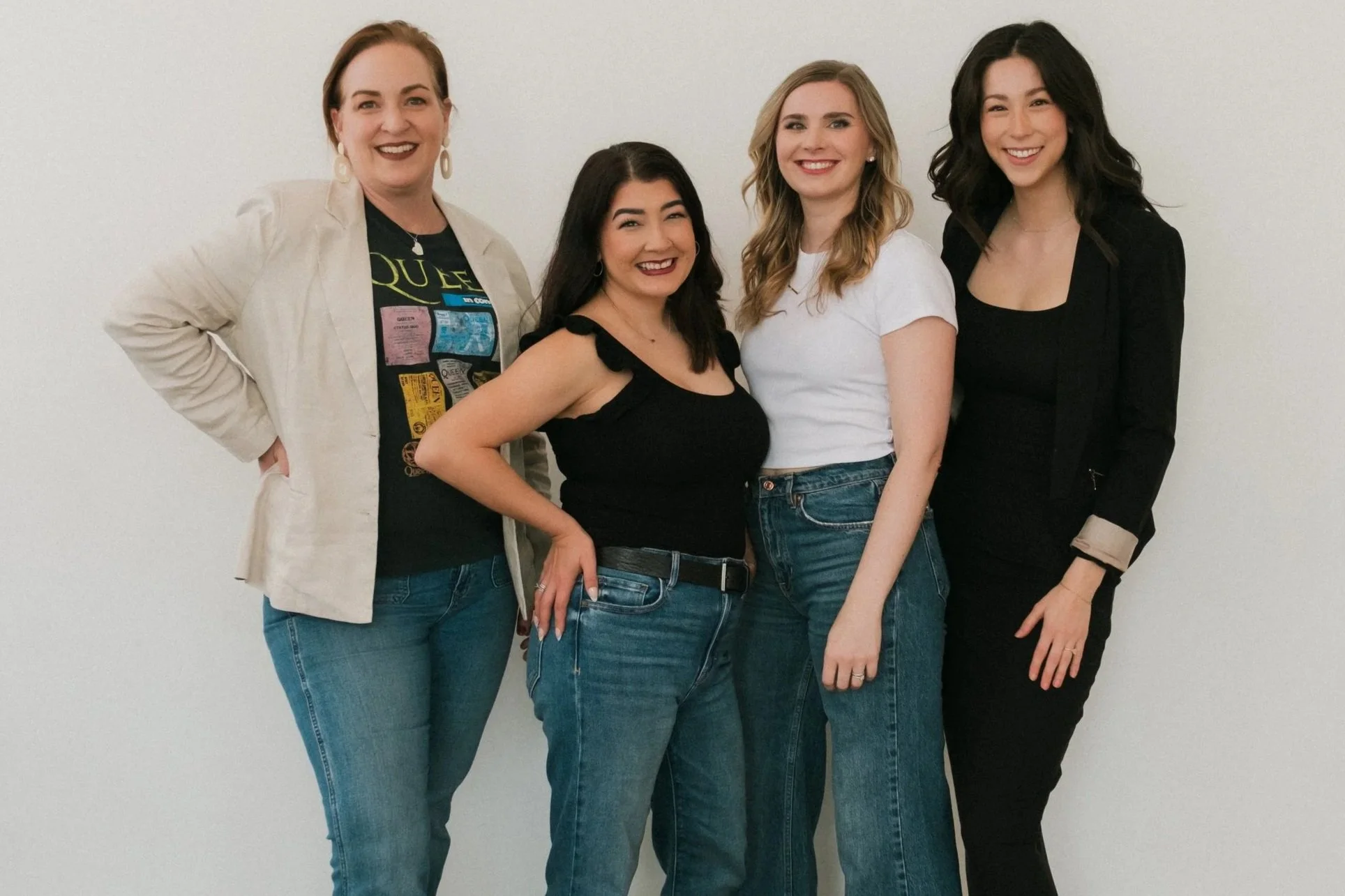Five women standing against a plain white wall, smiling and wearing casual clothes, with four women wearing jeans and all in different colored tops and blazers.