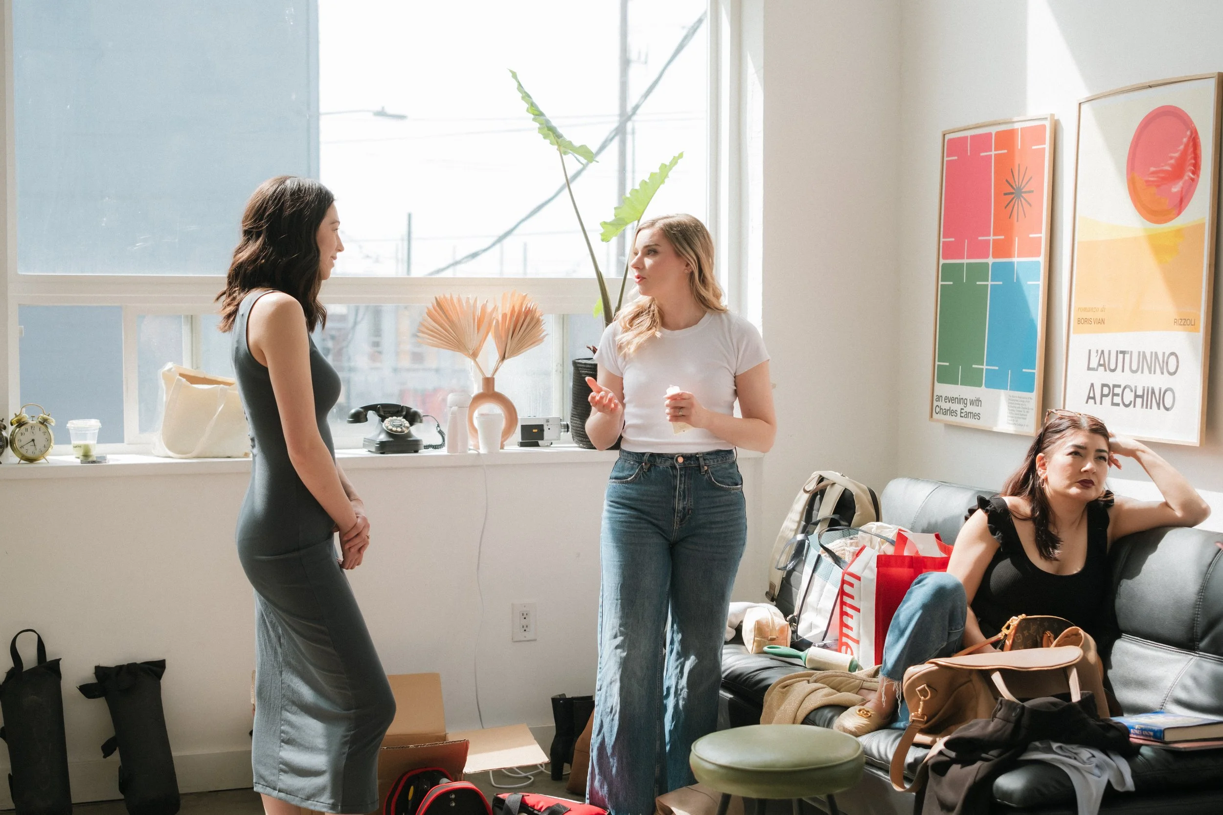 Two women are standing and talking in a bright room with large windows. One woman has dark hair and is wearing a gray dress, while the other has blonde hair and is dressed in a white T-shirt and blue jeans. There are several bags, personal items, and shopping bags on a black couch and the floor. The wall behind them has colorful artwork with geometric shapes and text.