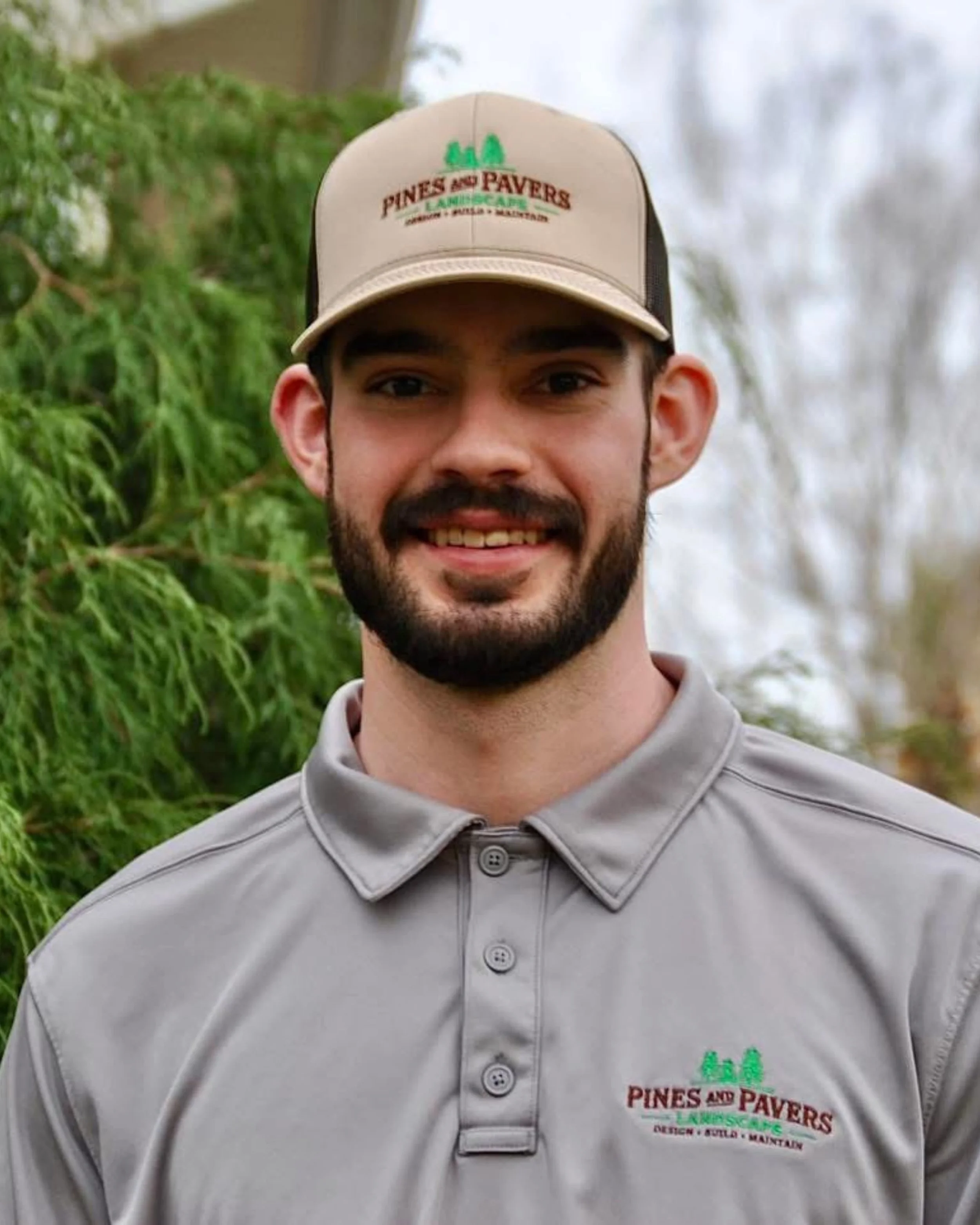A smiling man with dark hair and a beard wearing a beige cap and a gray polo shirt with a logo that says 'Pines and Pavers Landscape' with trees on it, outdoors with green foliage and trees in the background.