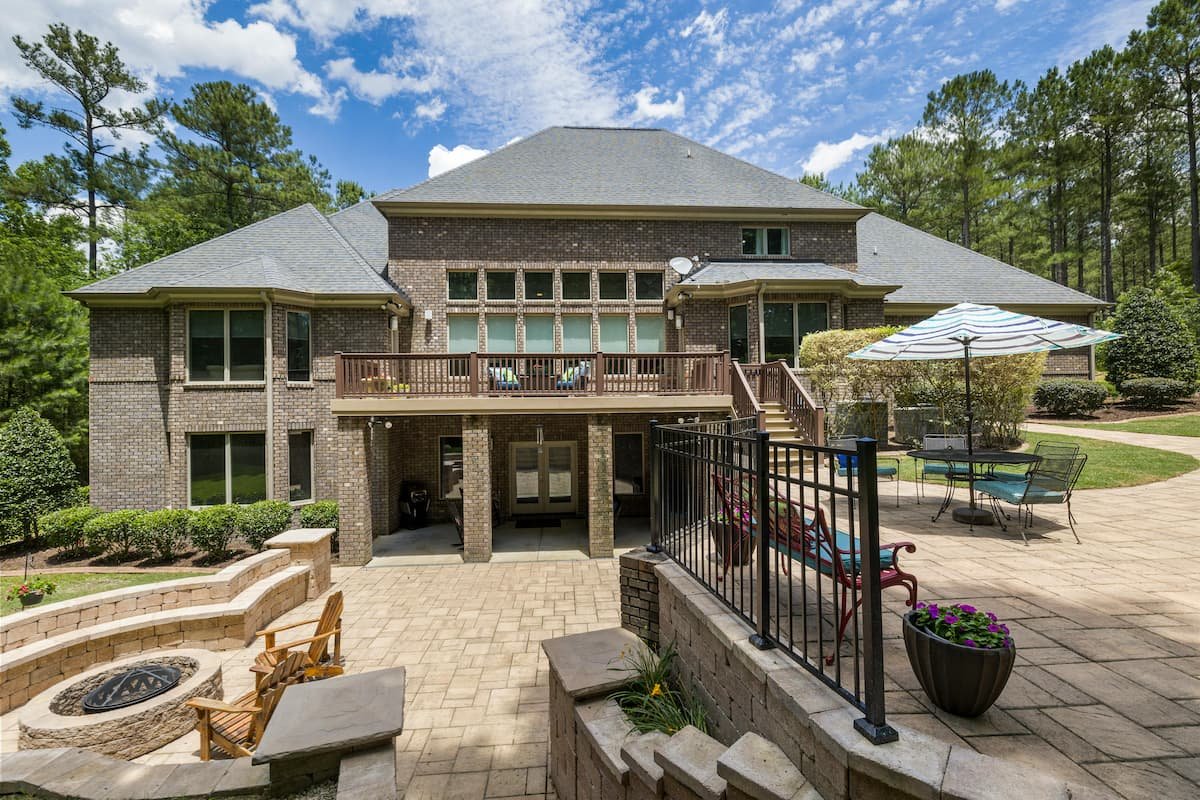 Large luxury backyard patio featuring layered paver hardscaping, fire pit seating area, retaining walls, and elevated deck overlooking a wooded property.