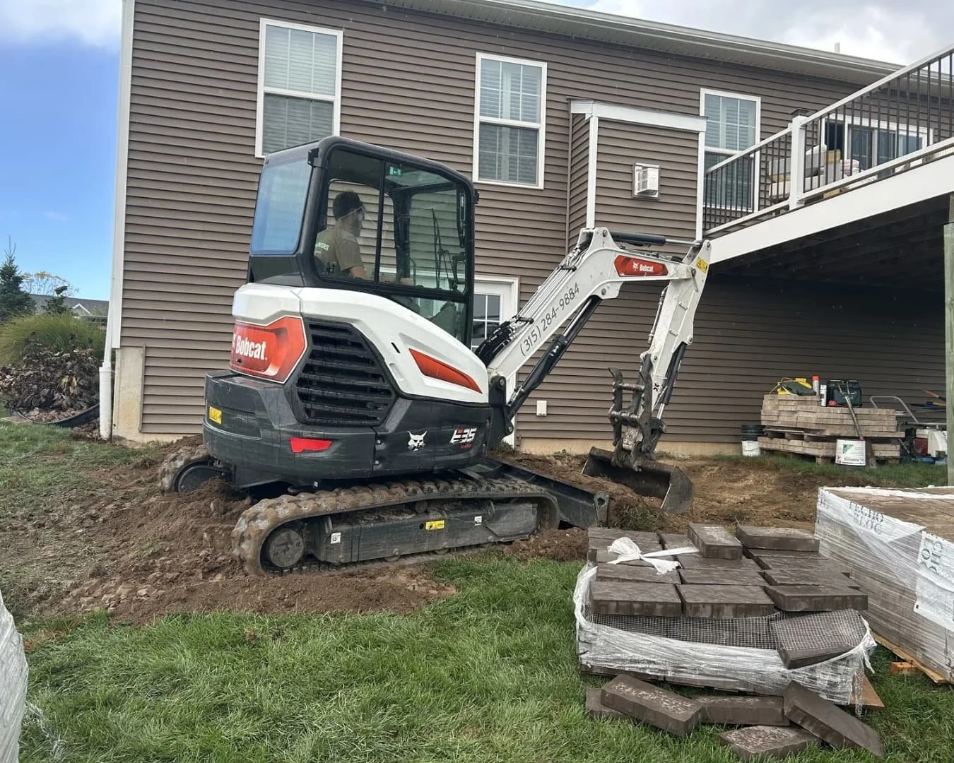 A Bobcat mini excavator working on a construction site near a residential building with brown siding, a deck, and stacked pavers in the foreground.