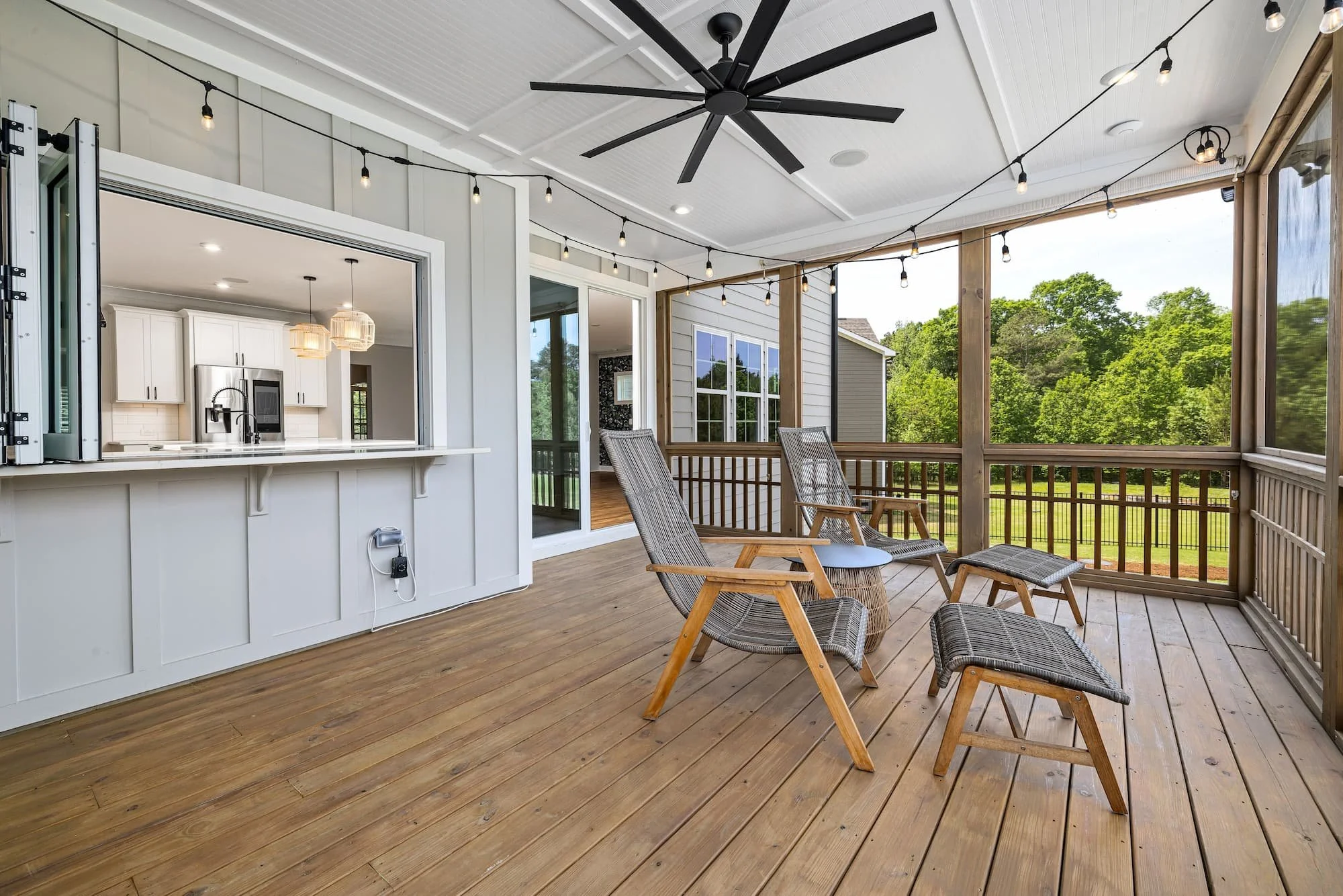 Spacious screened porch featuring warm wood decking, a pass-through kitchen window, string lights, ceiling fan, and comfortable seating designed for relaxing and entertaining outdoors.