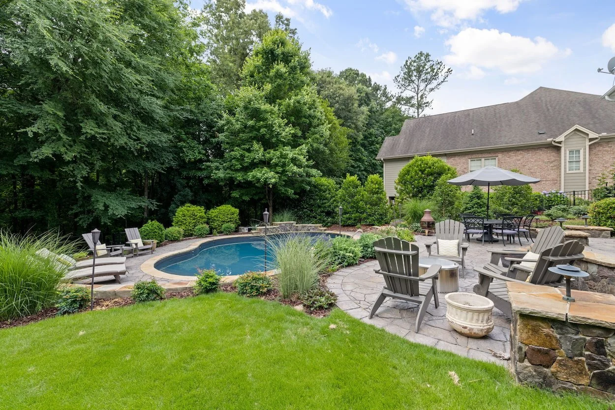 Beautiful backyard featuring a swimming pool, natural stone paver patio, Adirondack chairs, lush landscaping, and multiple gathering areas designed for relaxation and entertaining.