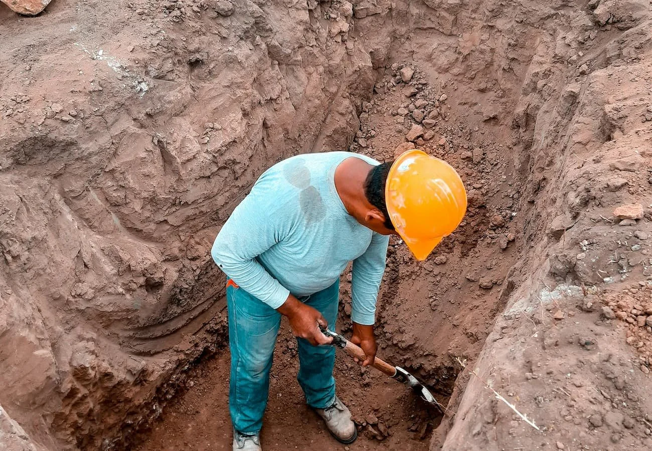 Construction worker digging a deep trench during landscape or hardscape foundation preparation with exposed soil and safety helmet.