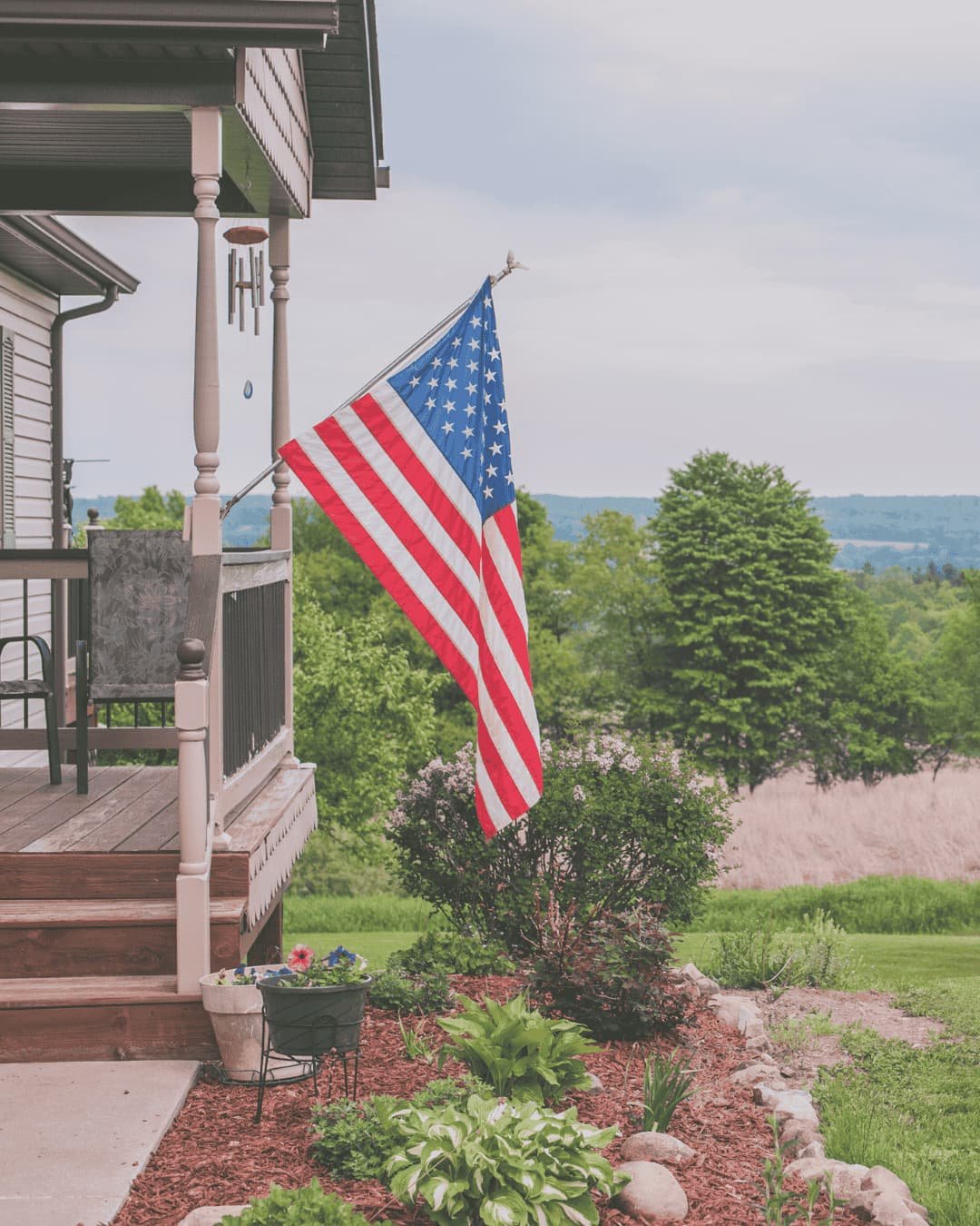 Front porch with American flag displayed above a freshly mulched garden bed featuring hostas, flowering shrubs, and natural stone edging overlooking a green lawn and rolling hills.