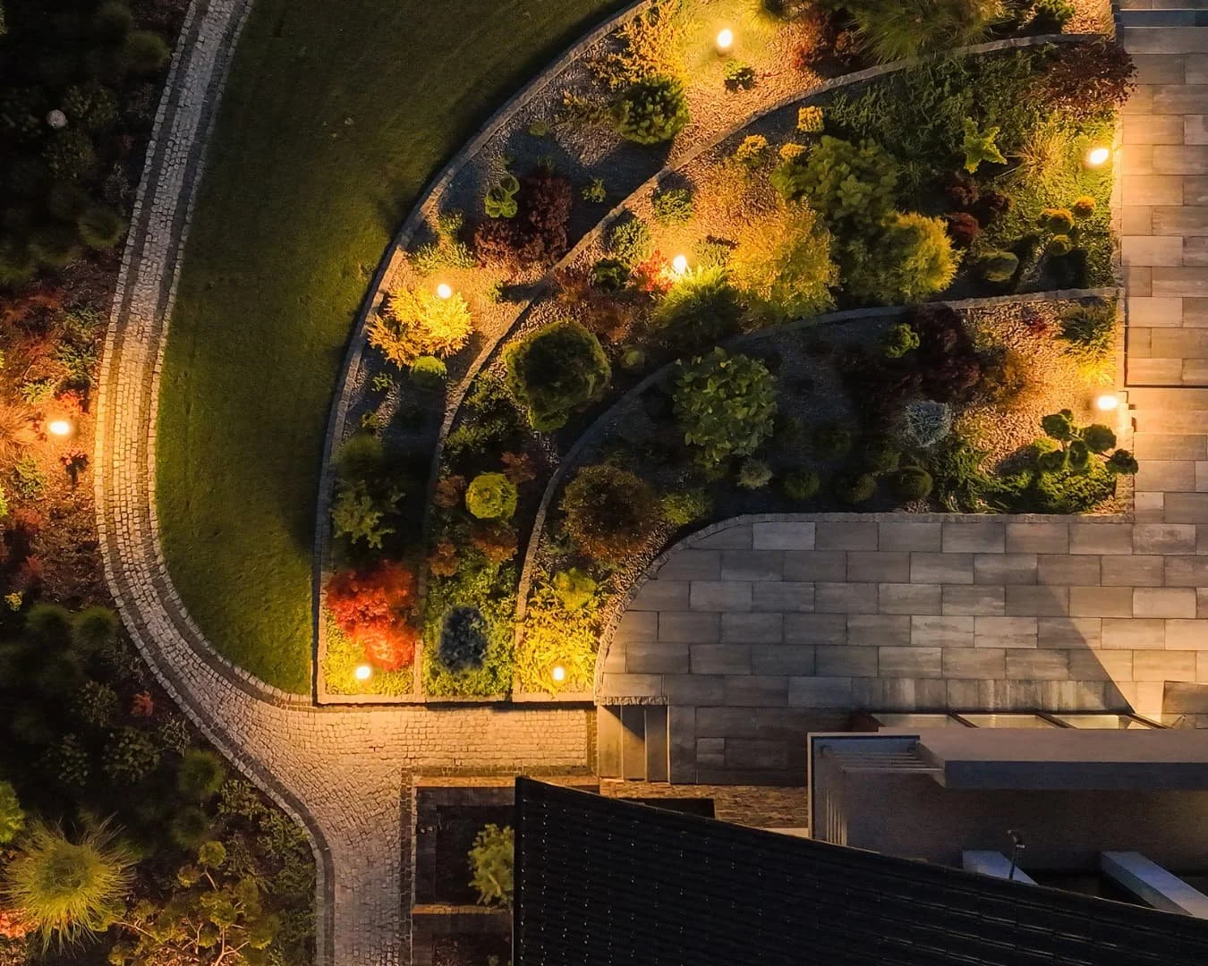 Outdoor lighting inspiration with warm overhead lighting illuminating a patio and planting beds at night.