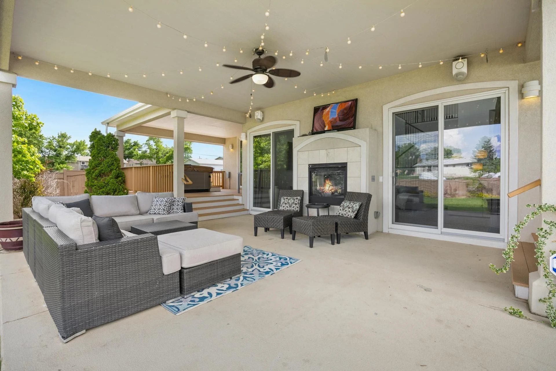 Outdoor patio with wicker sofa, chairs, fireplace, ceiling fan, string lights, and a tv, overlooking a backyard with trees and a fence.