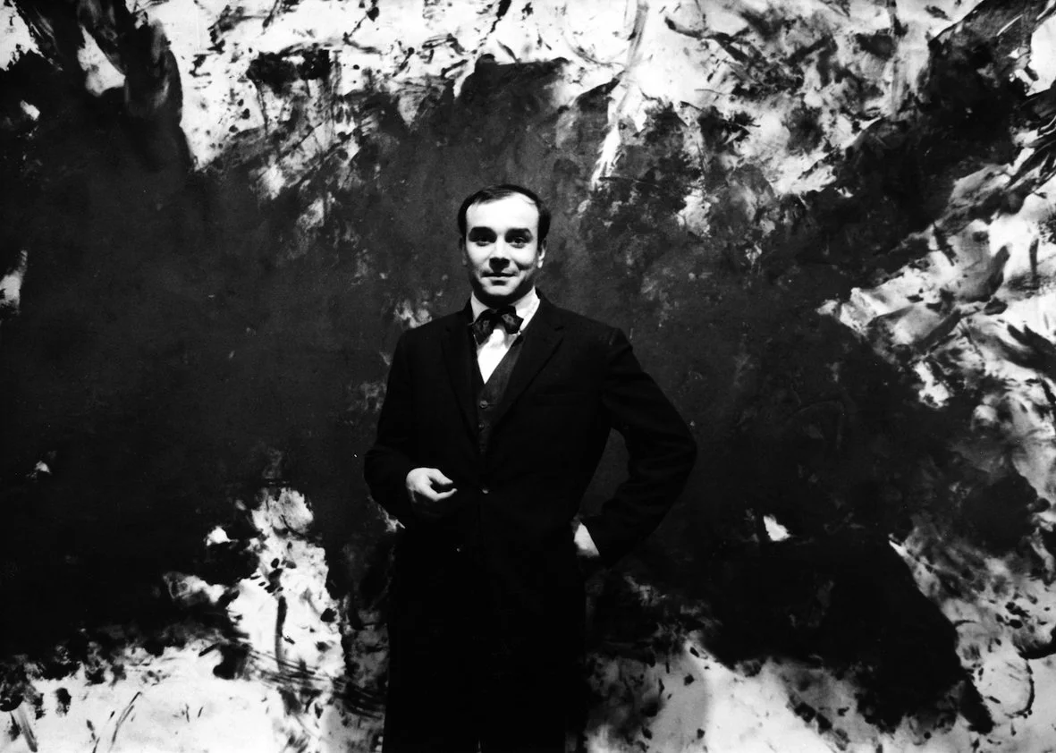 Yves Klein wearing a suit standing in front of an abstract black and white painting.