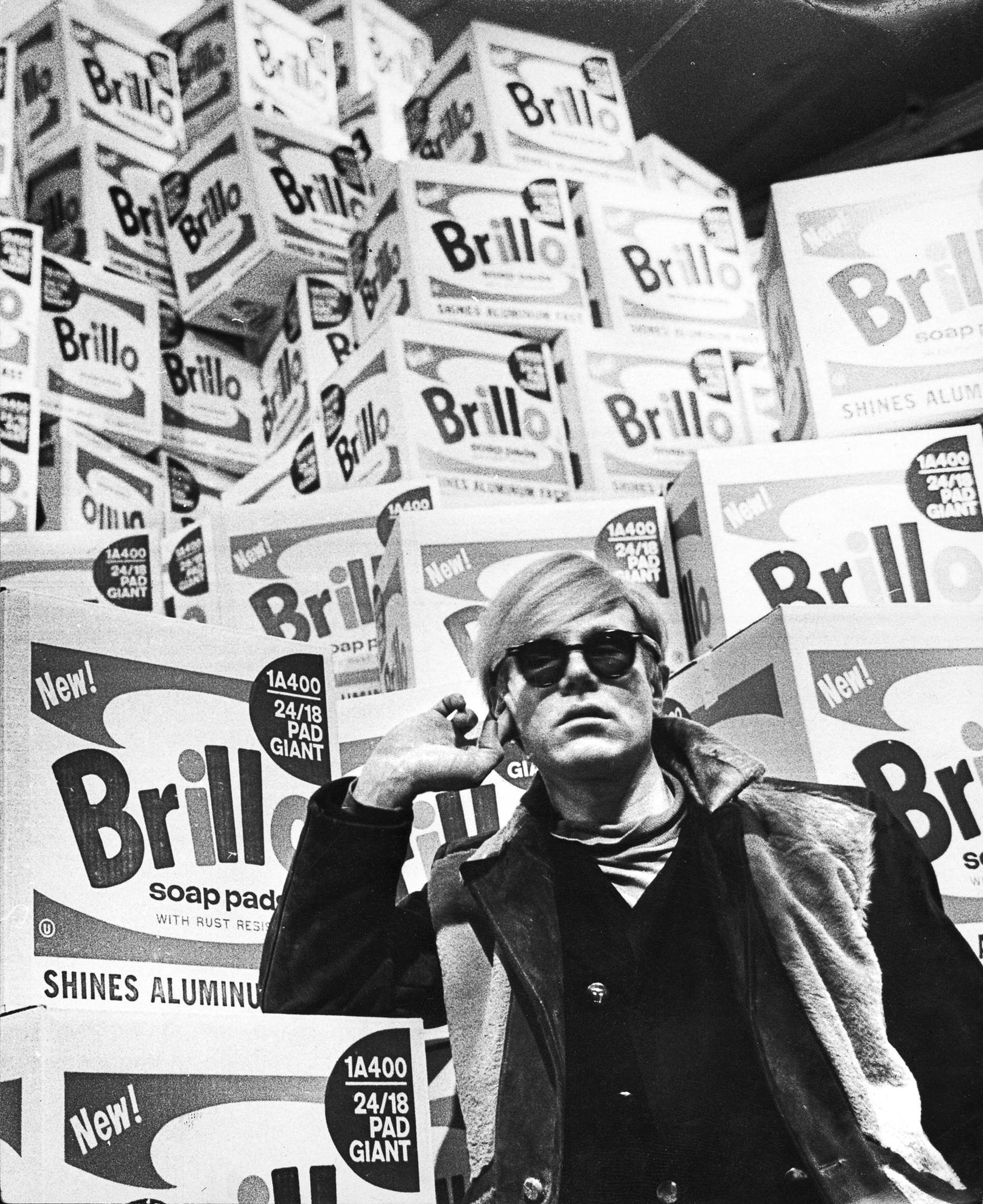 Andy Warhol posing infront of Brillo boxes