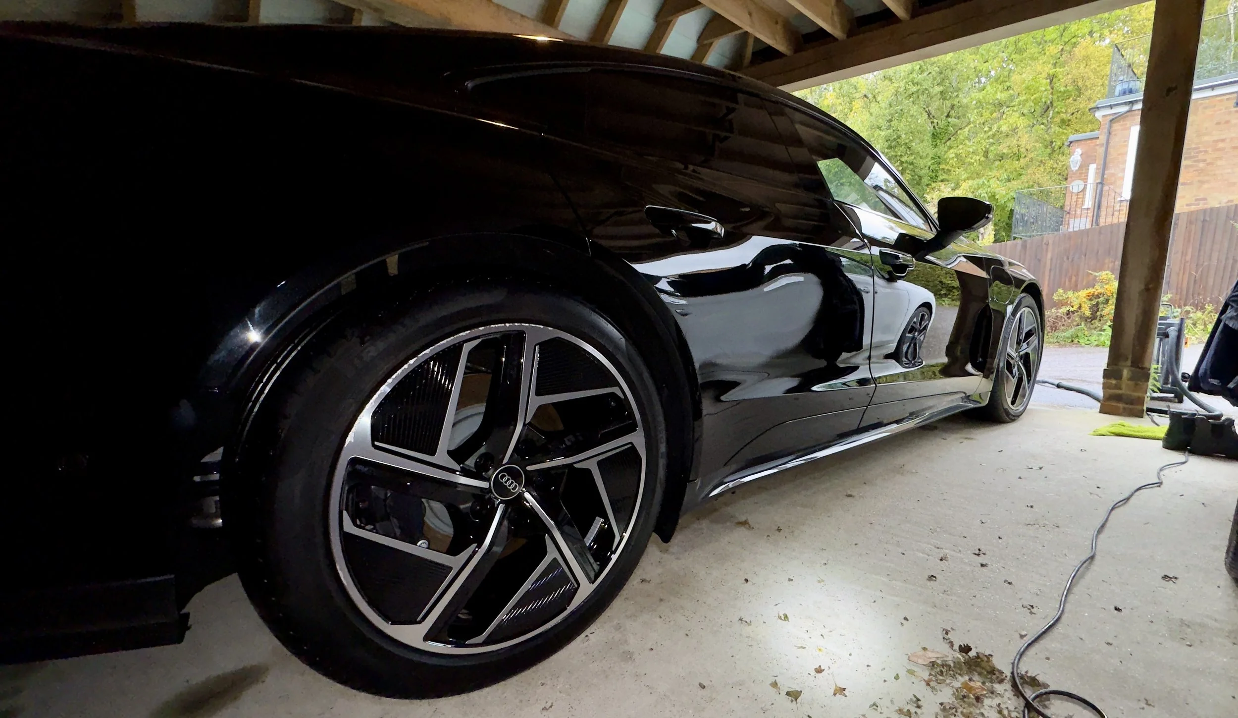 A black Audi sports car parked in a garage with a wooden roof and open side revealing a residential backyard with trees and a brick house.